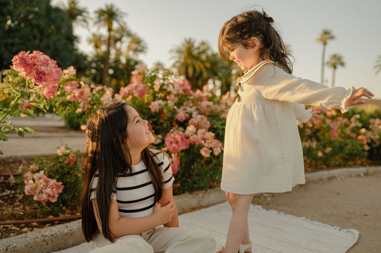 A little girl twirls in a white dress while her sister watches from a rose garden, a family session in Glendale, AZ.