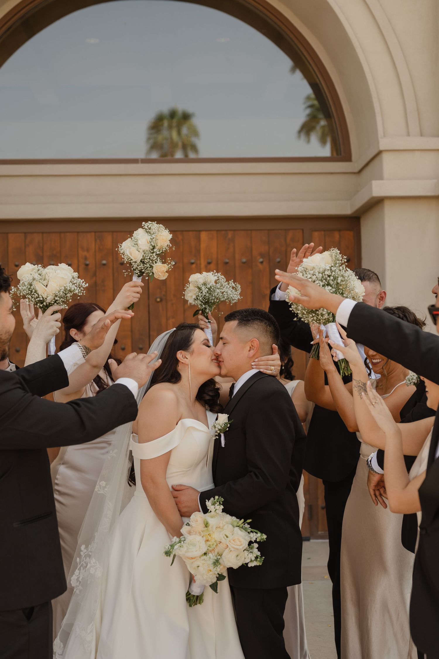 Wedding party cheering as bride and groom kiss under floral arch, joyful group celebration outside ceremony venue.