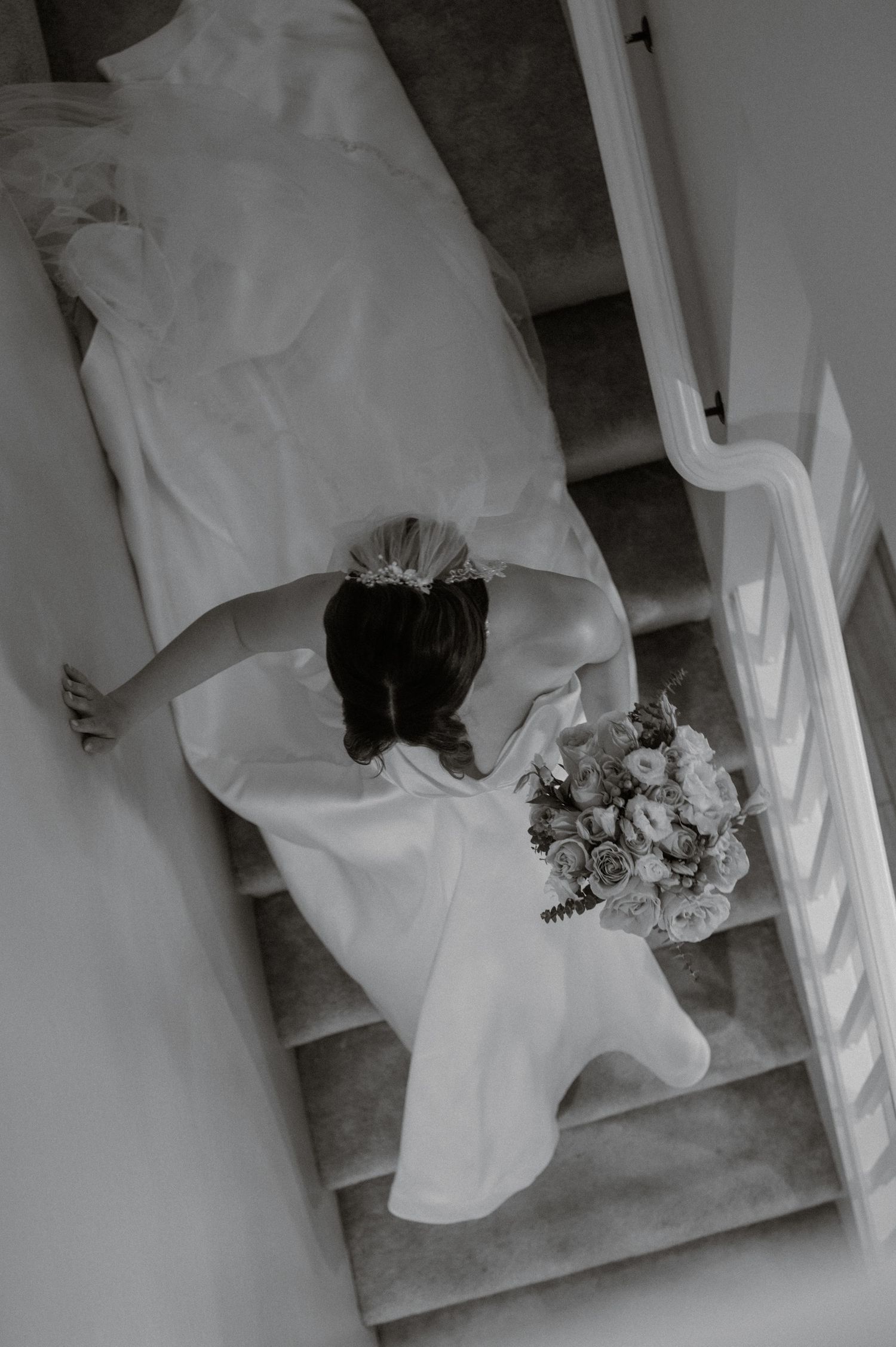 Overhead black-and-white shot of bride descending staircase with long train and bouquet in hand.