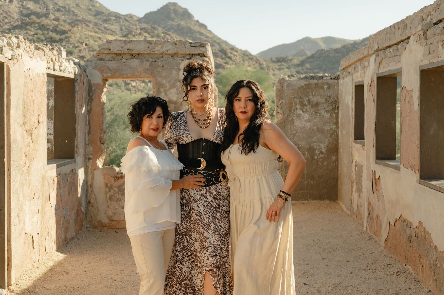 Three women pose in flowing attire before rustic stone ruins with mountain backdrop in desert setting.