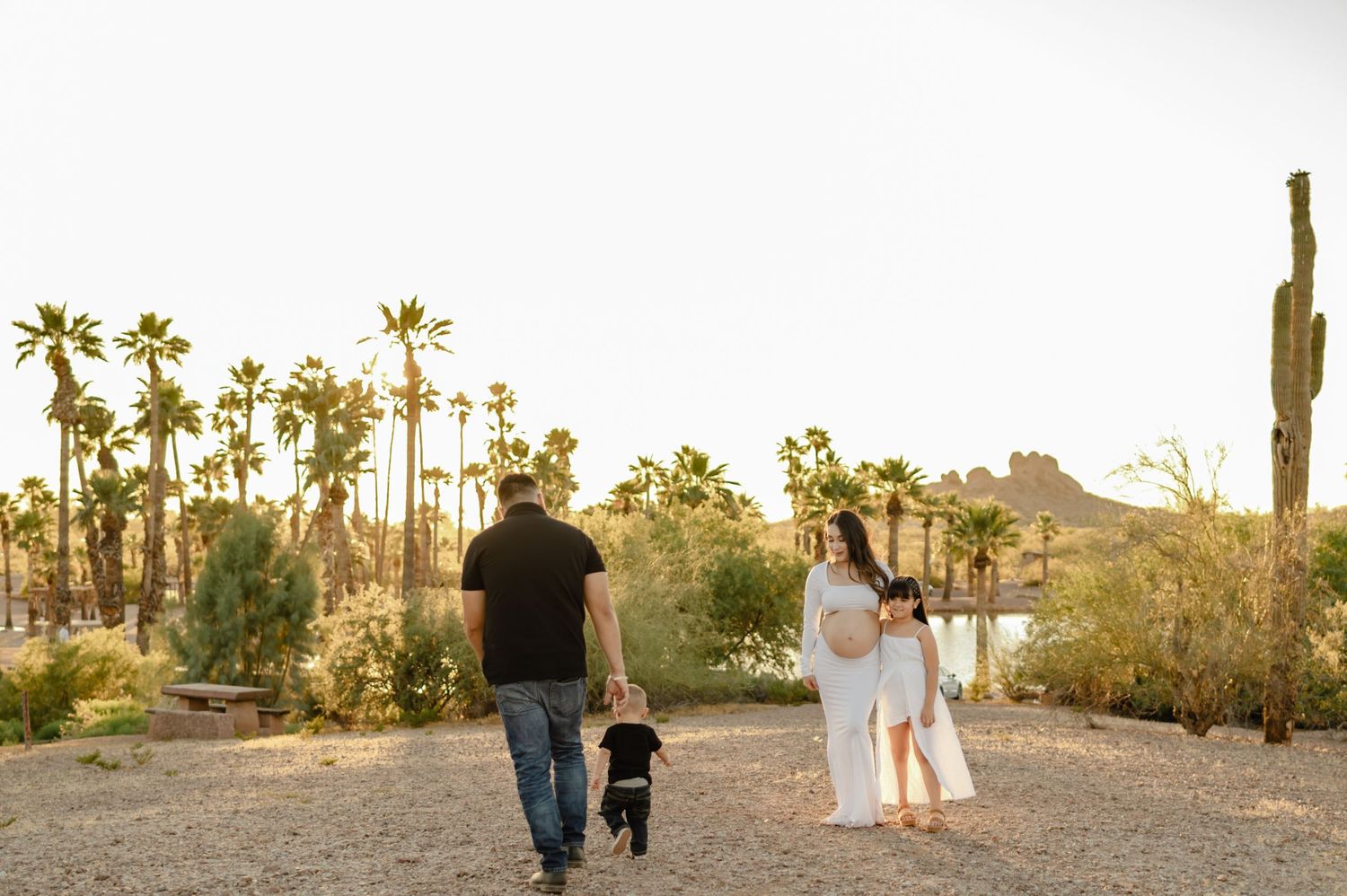 Family walking together in a desert landscape with palm trees and saguaro cacti during golden hour maternity photo session.