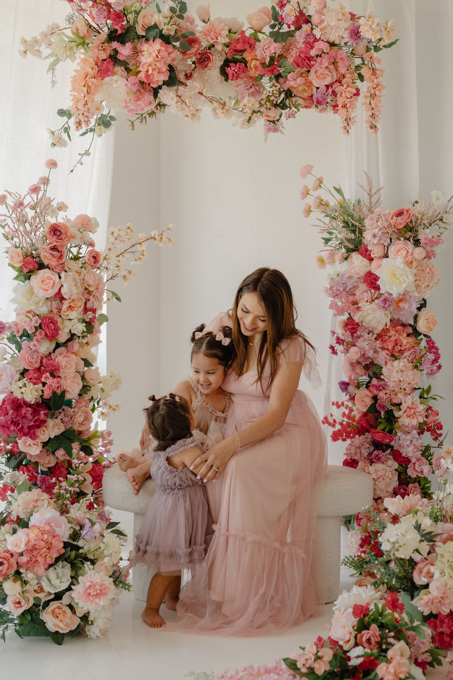 A mother in pink poses with her two daughters under a floral arch during a mommy and me mini at Light Loft in Scottsdale, AZ.