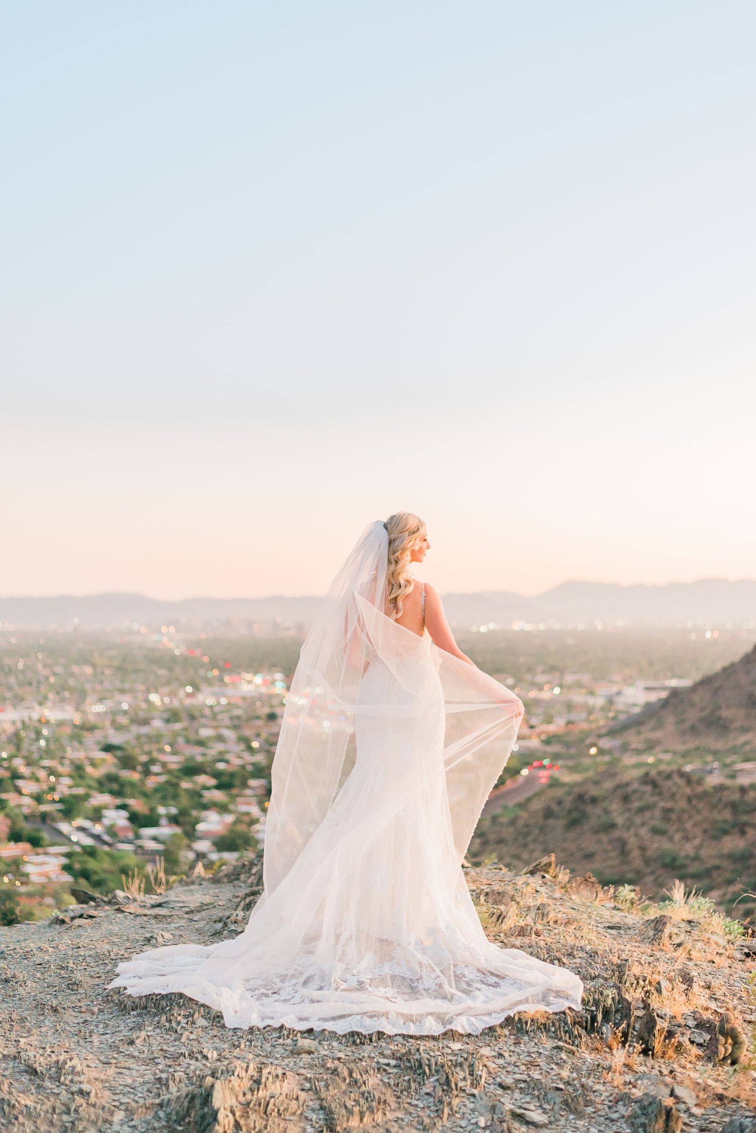 Bride stands on hilltop at sunset overlooking panoramic desert cityscape.