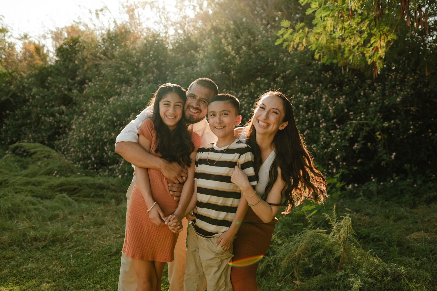 family of four embraces outdoors surrounded by lush greenery during golden hour in mesa, arizona
