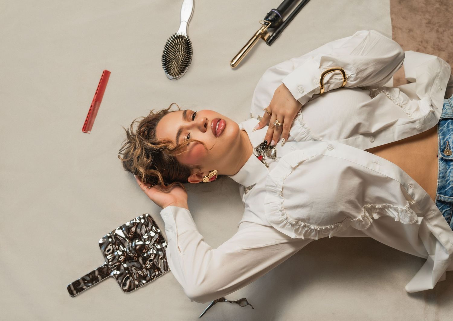 Woman lying on the floor surrounded by curling irons and brushes, holding a mascara wand to her lashes — part of The Hive Salon shoot.