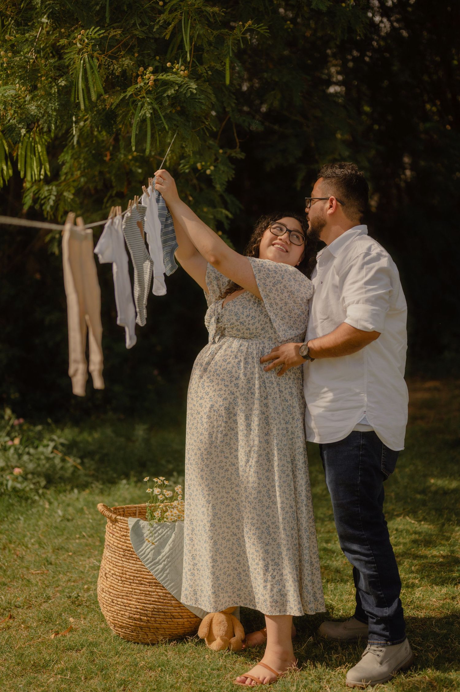 Pregnant woman and husband hanging tiny baby clothes on line in lush garden at golden hour.