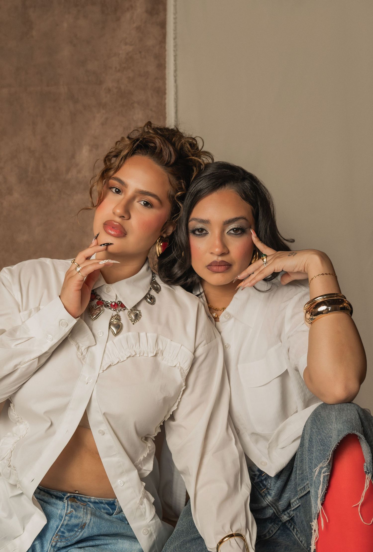 Two women sit closely with playful expressions in front of a tan backdrop.
