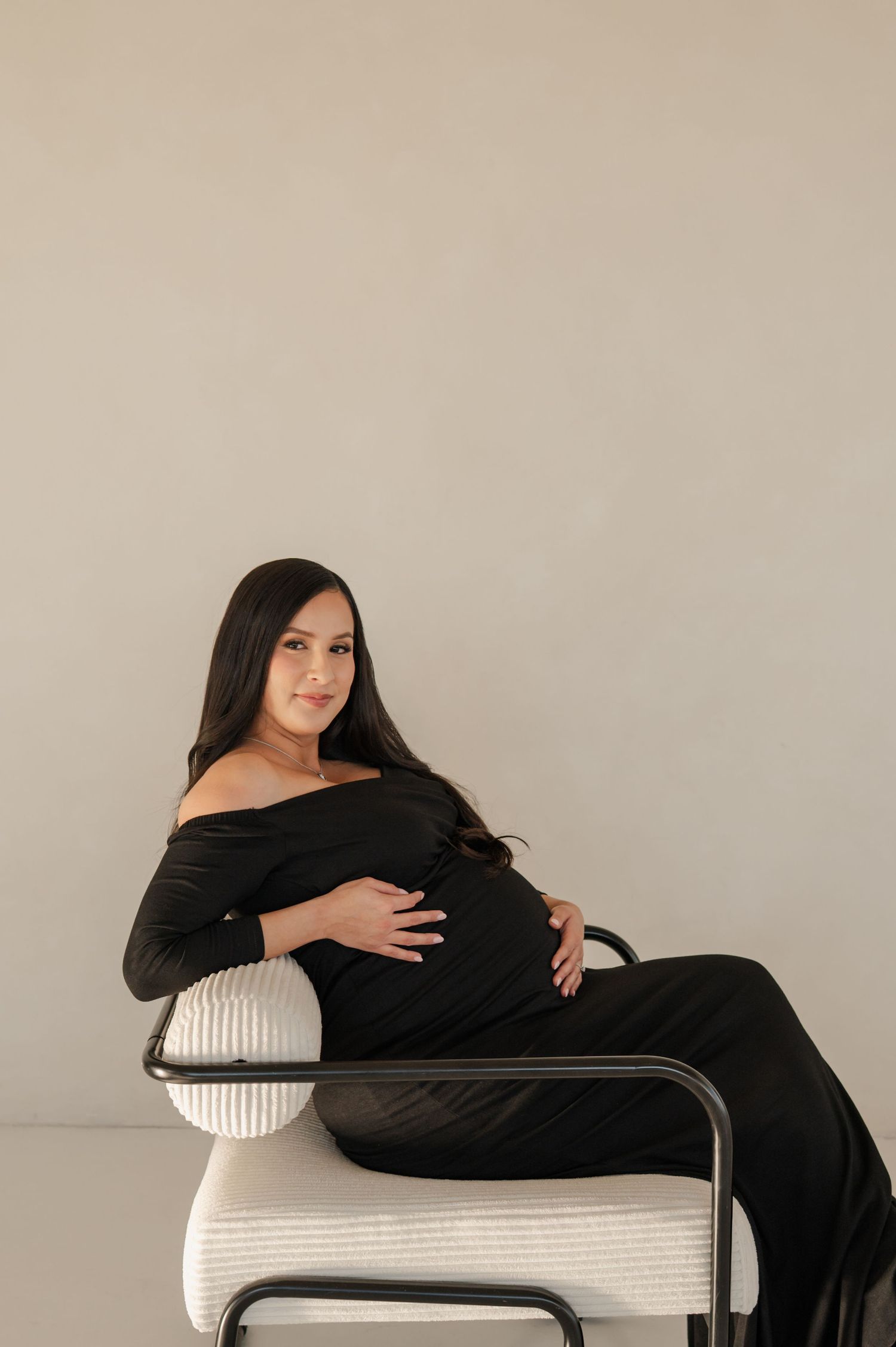 pregnant woman in a black dress sits in a chair cradling her bump during a phoenix studio session