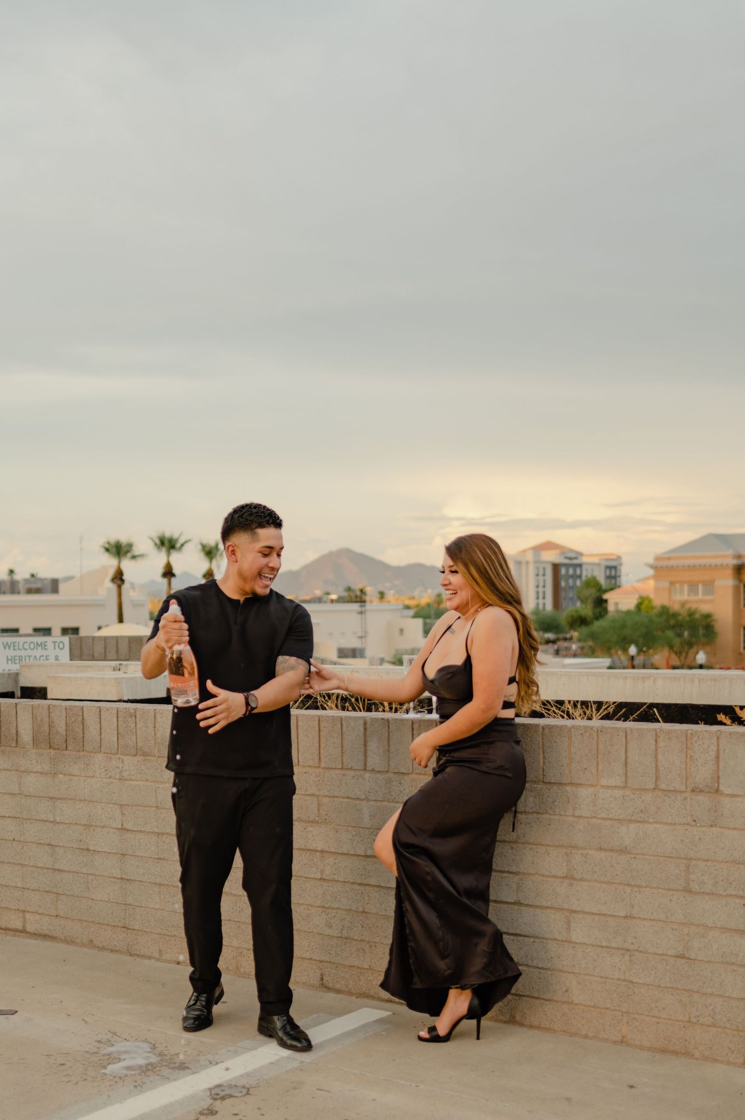 Couple in black formal attire sharing a champagne bottle on a rooftop at sunset with mountains in background.