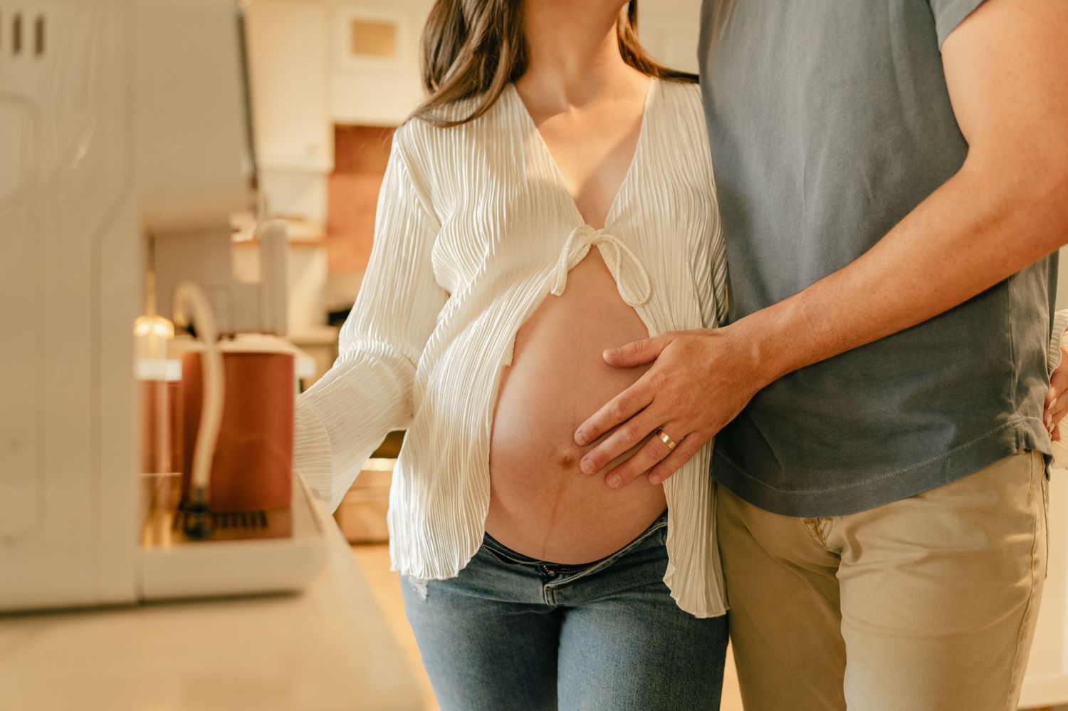 A pregnant woman in a white blouse and jeans embraces her baby bump with her partner during an in home maternity session.
