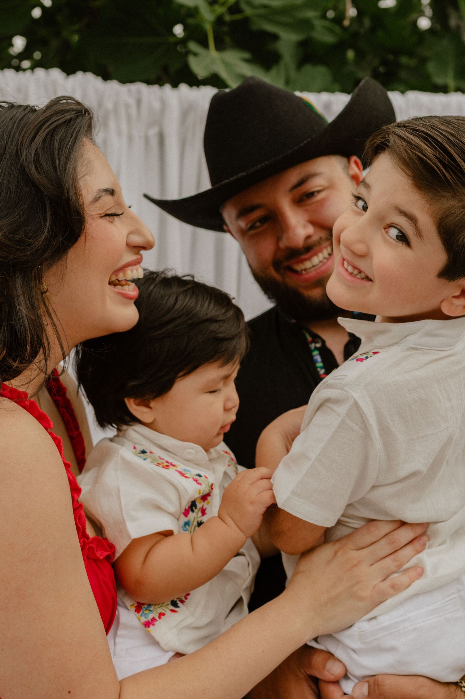 Happy family of four laughing together outdoors, father wearing black cowboy hat.