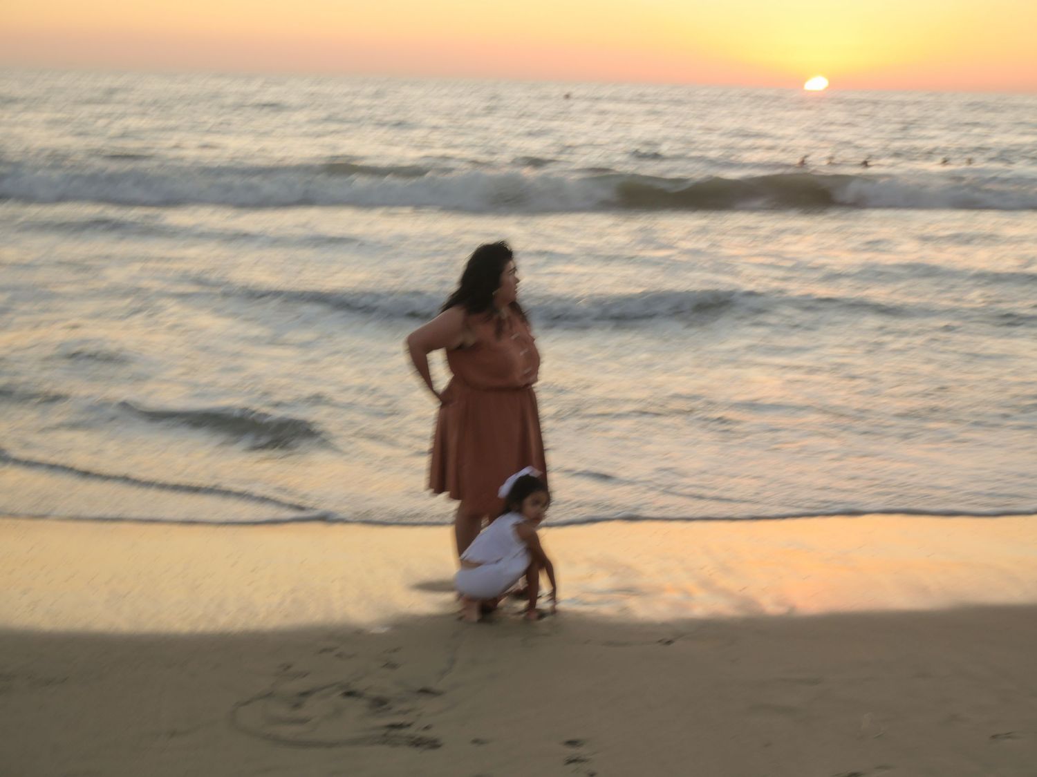 Woman in orange dress standing on beach at sunset with child playing in sand near ocean waves.