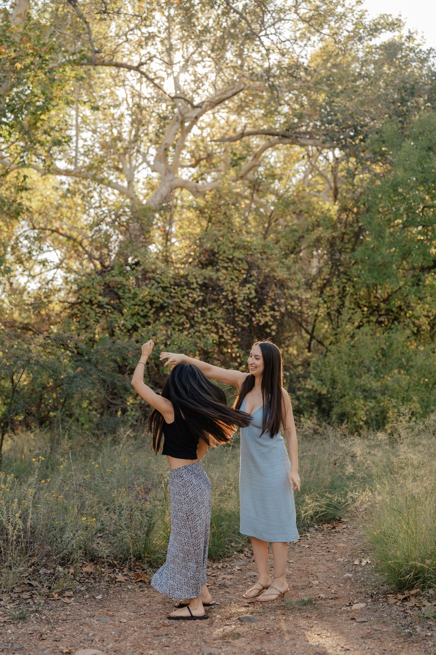 Mother and daughter duo laugh and dance together on a sunlit forest path, a candid portrait session.