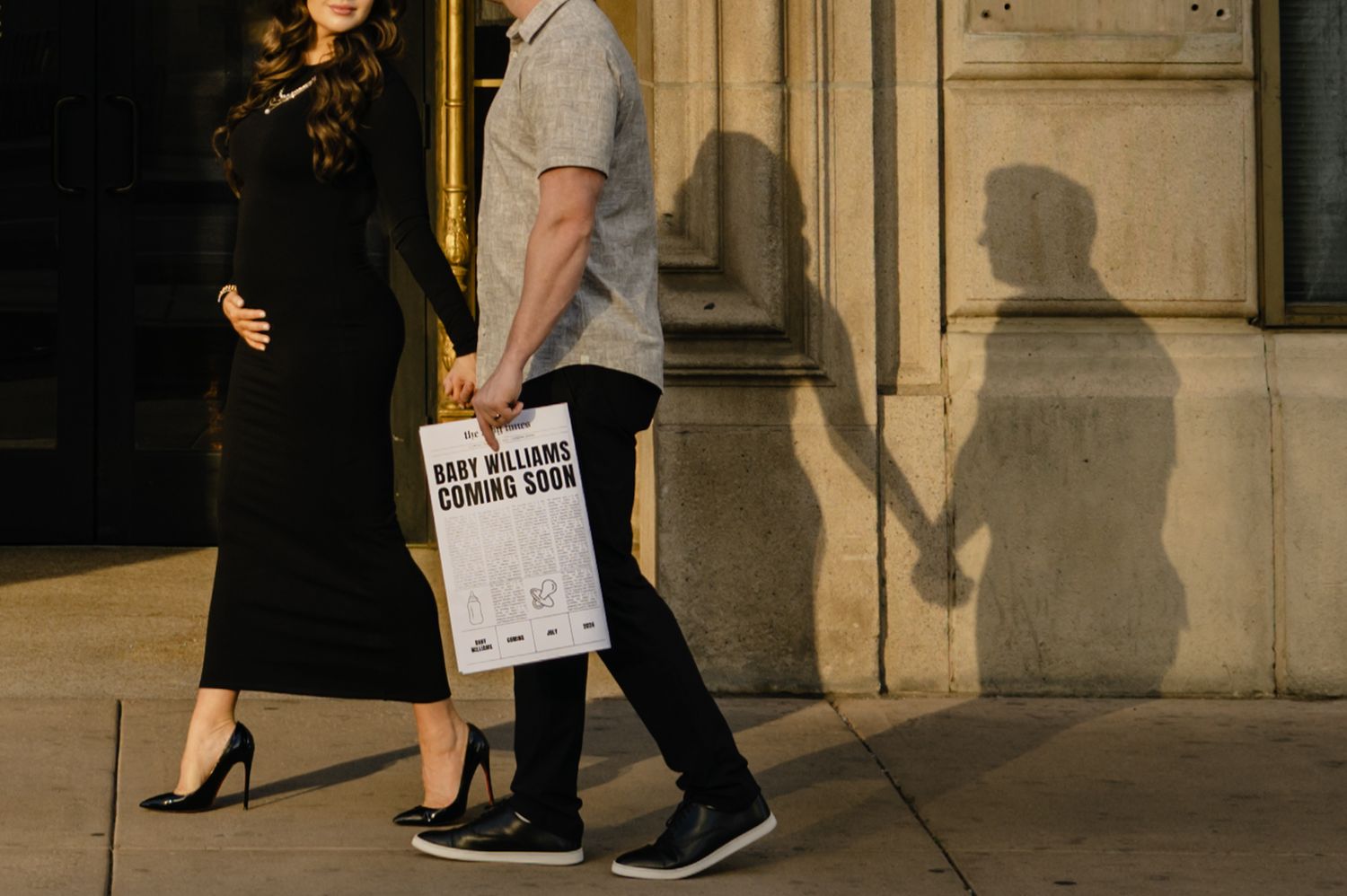 Couple holding hands and a baby announcement sign casting shadows on a building wall during a maternity photo shoot.