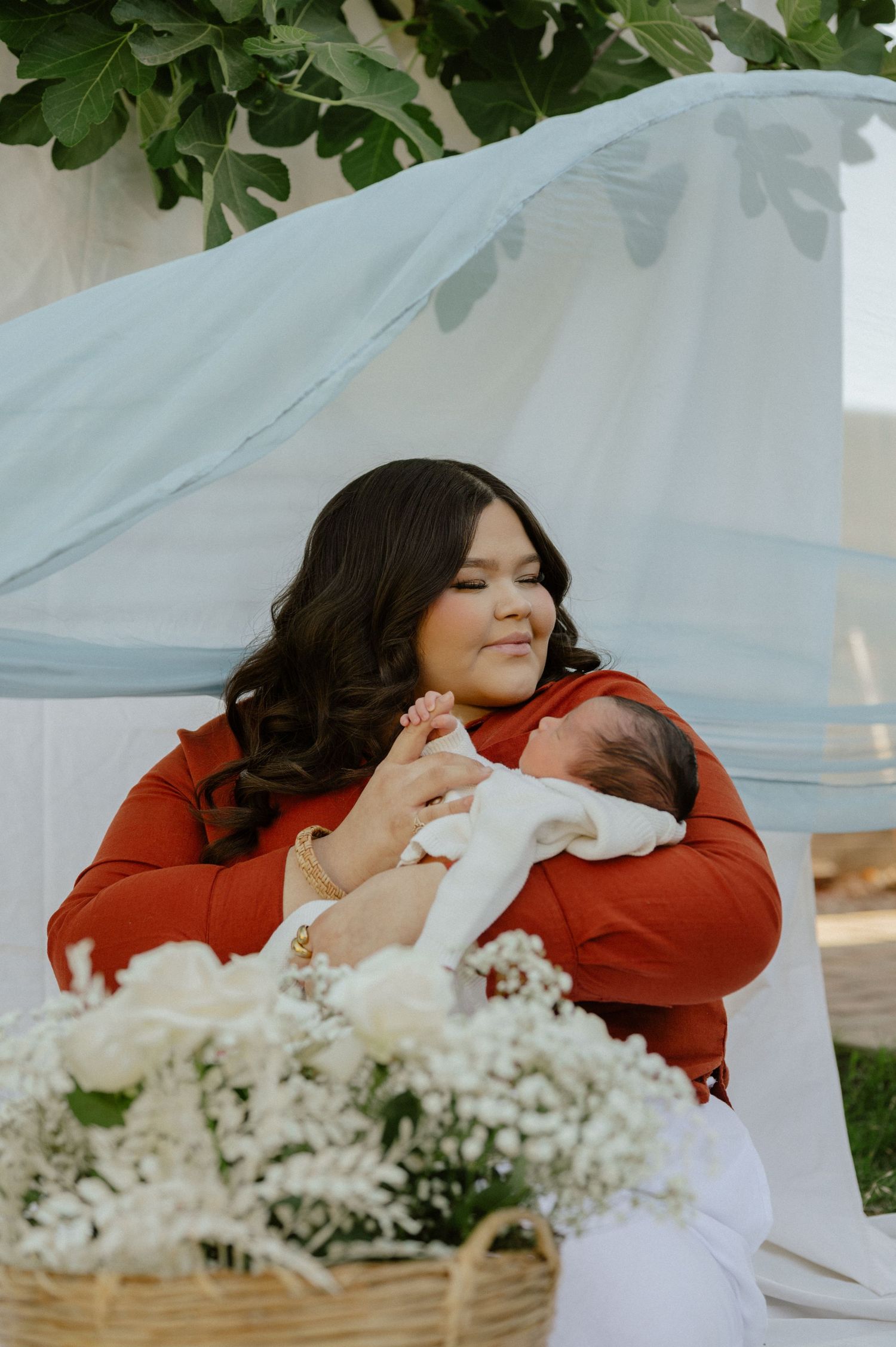 A woman in red cradles a sleeping newborn outdoors beneath a soft blue canopy of flowers.