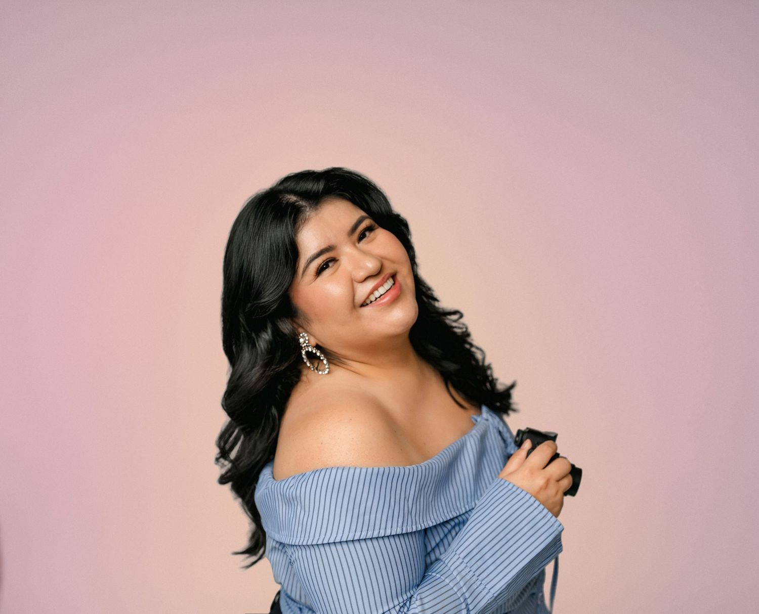 Smiling young woman with black hair wearing a blue off-shoulder striped top against a pink background.