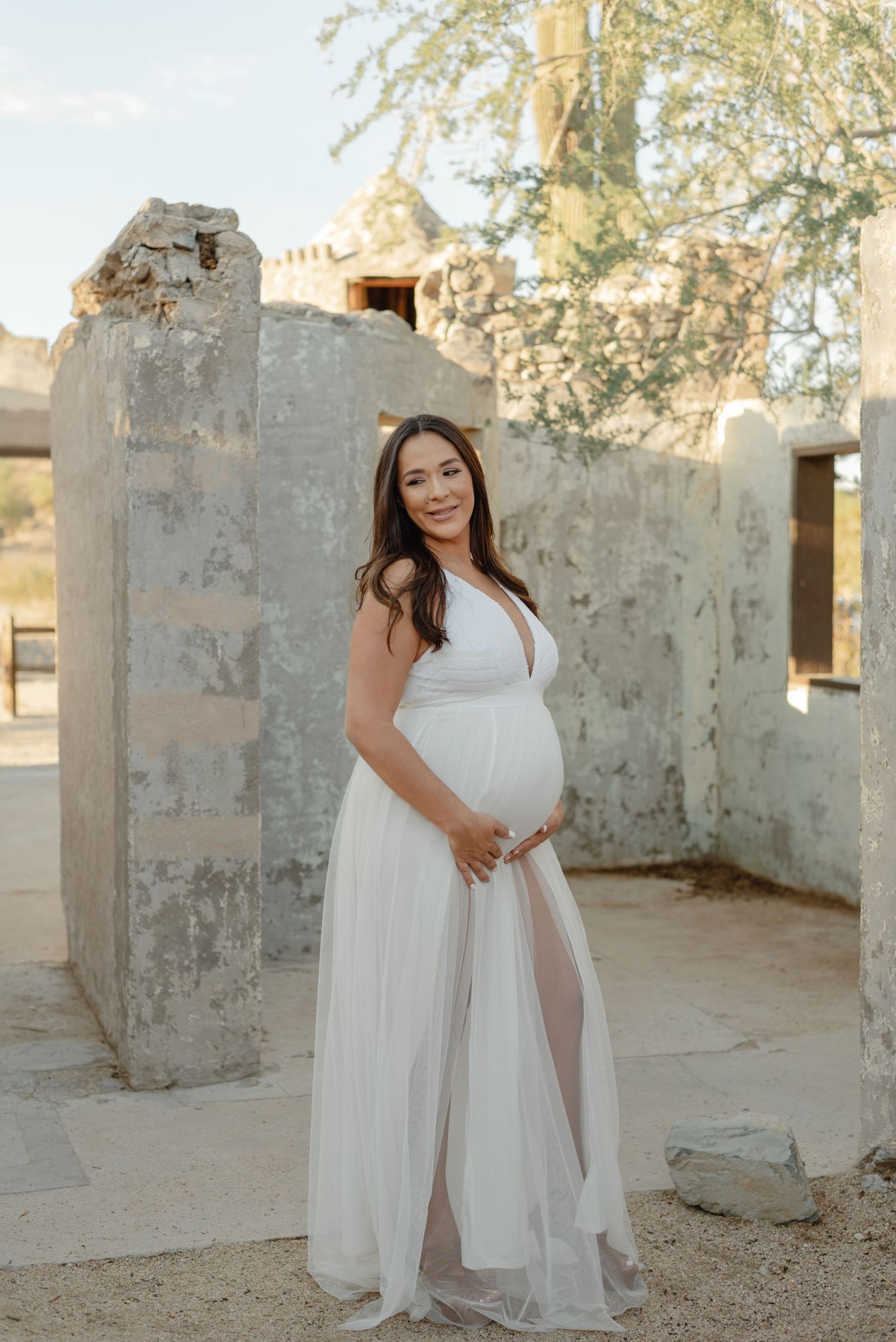 Smiling pregnant woman in white flowing gown cradling baby bump, standing between stone pillars outdoors.