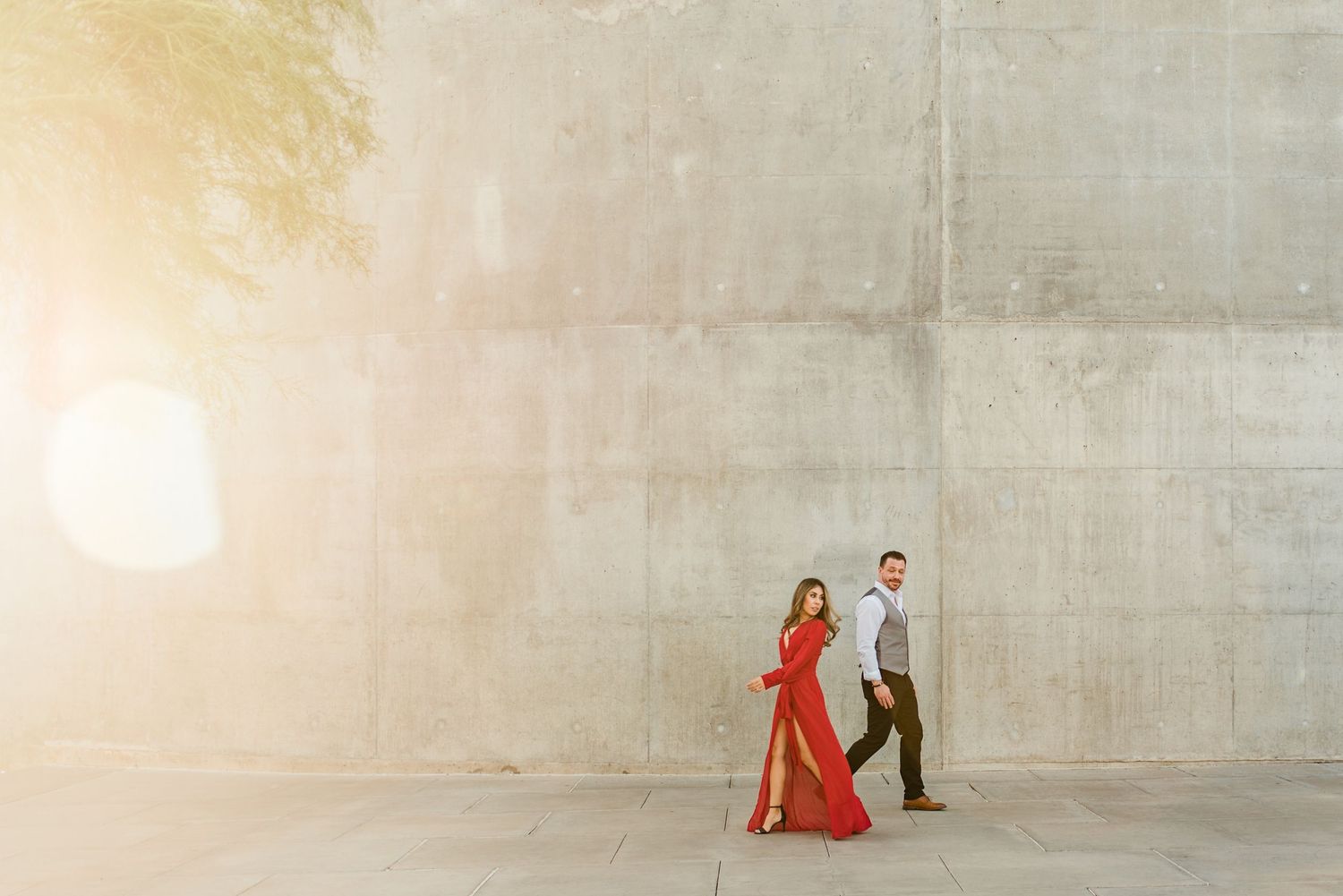 Couple in red dress and gray suit walking along concrete wall bathed in golden sunlight.