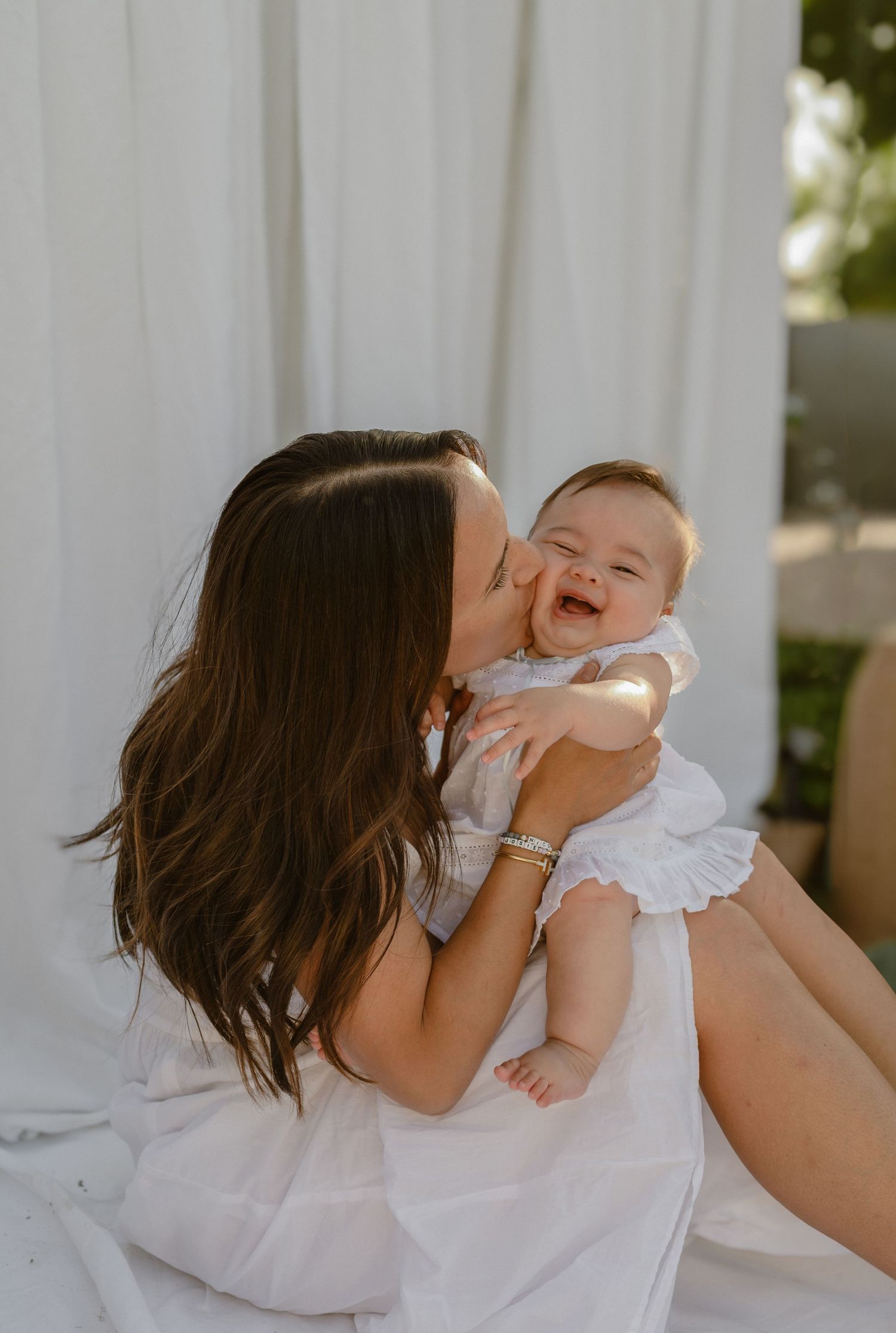 Mother kissing her laughing baby dressed in white during an outdoor portrait session with soft natural light.