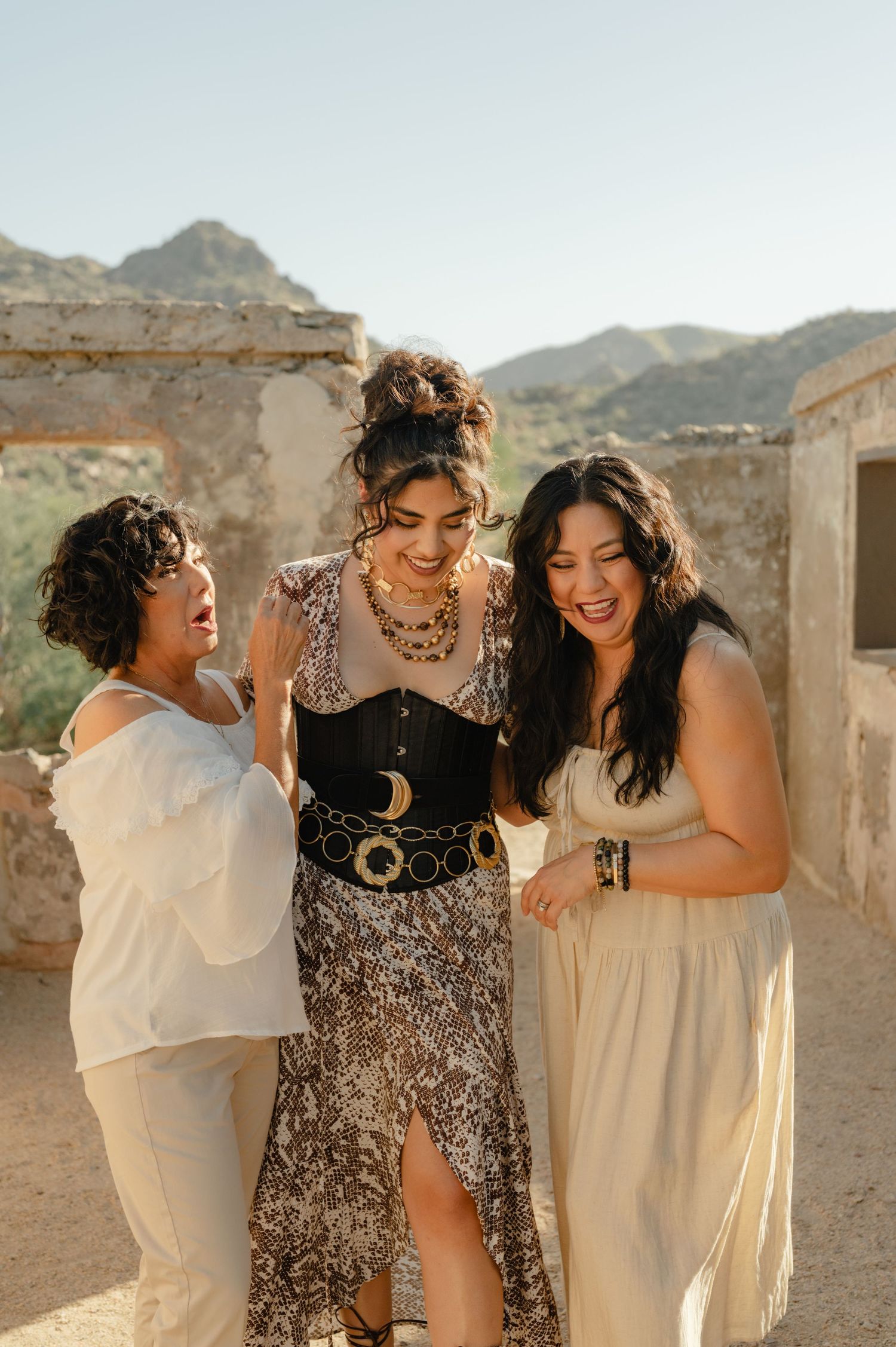 Three women smiling and embracing while dressed in desert-toned outfits, surrounded by sunlit ruins.