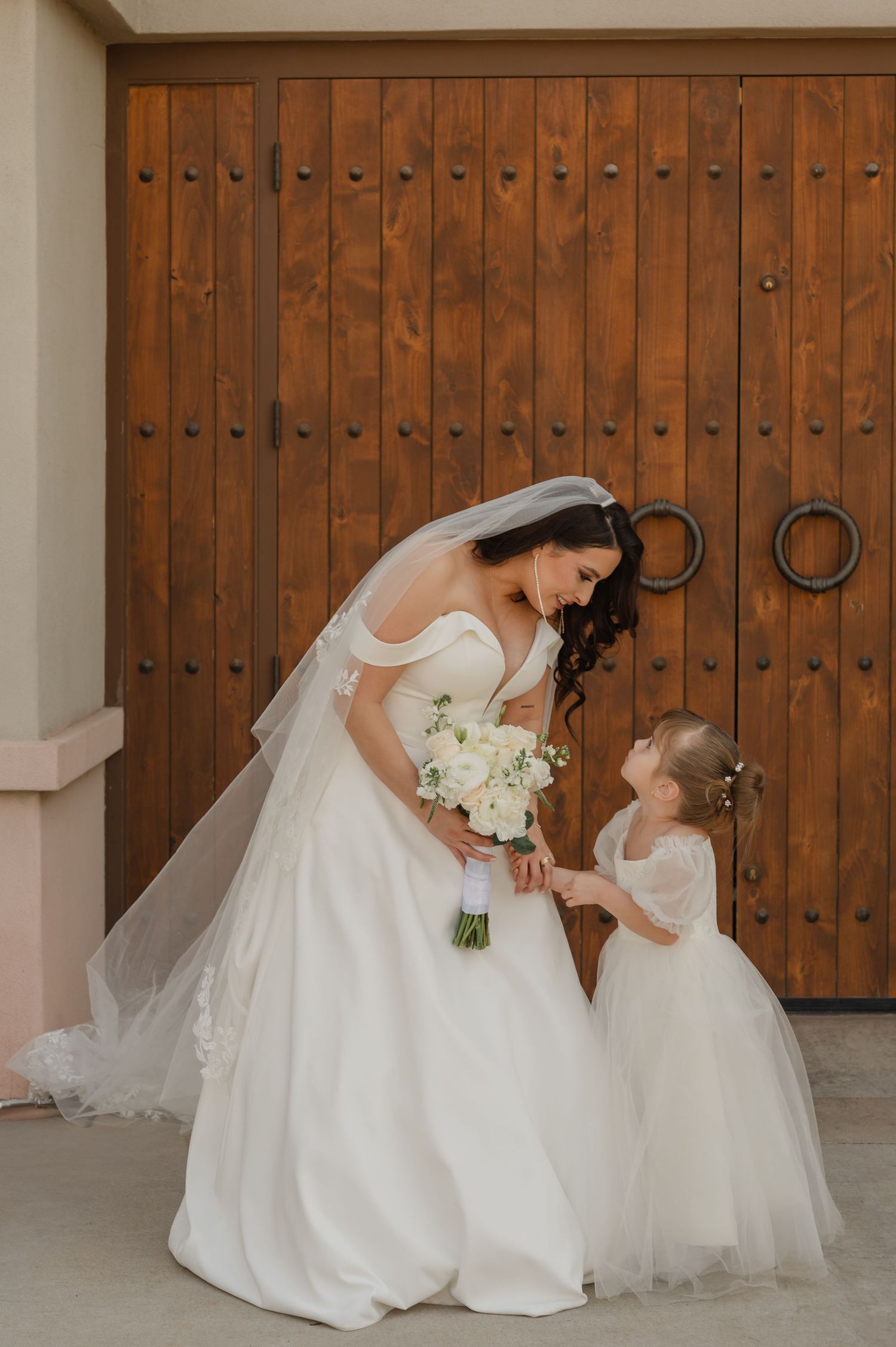 Bride kneeling to talk to flower girl in matching white dress, wooden doors in the background.
