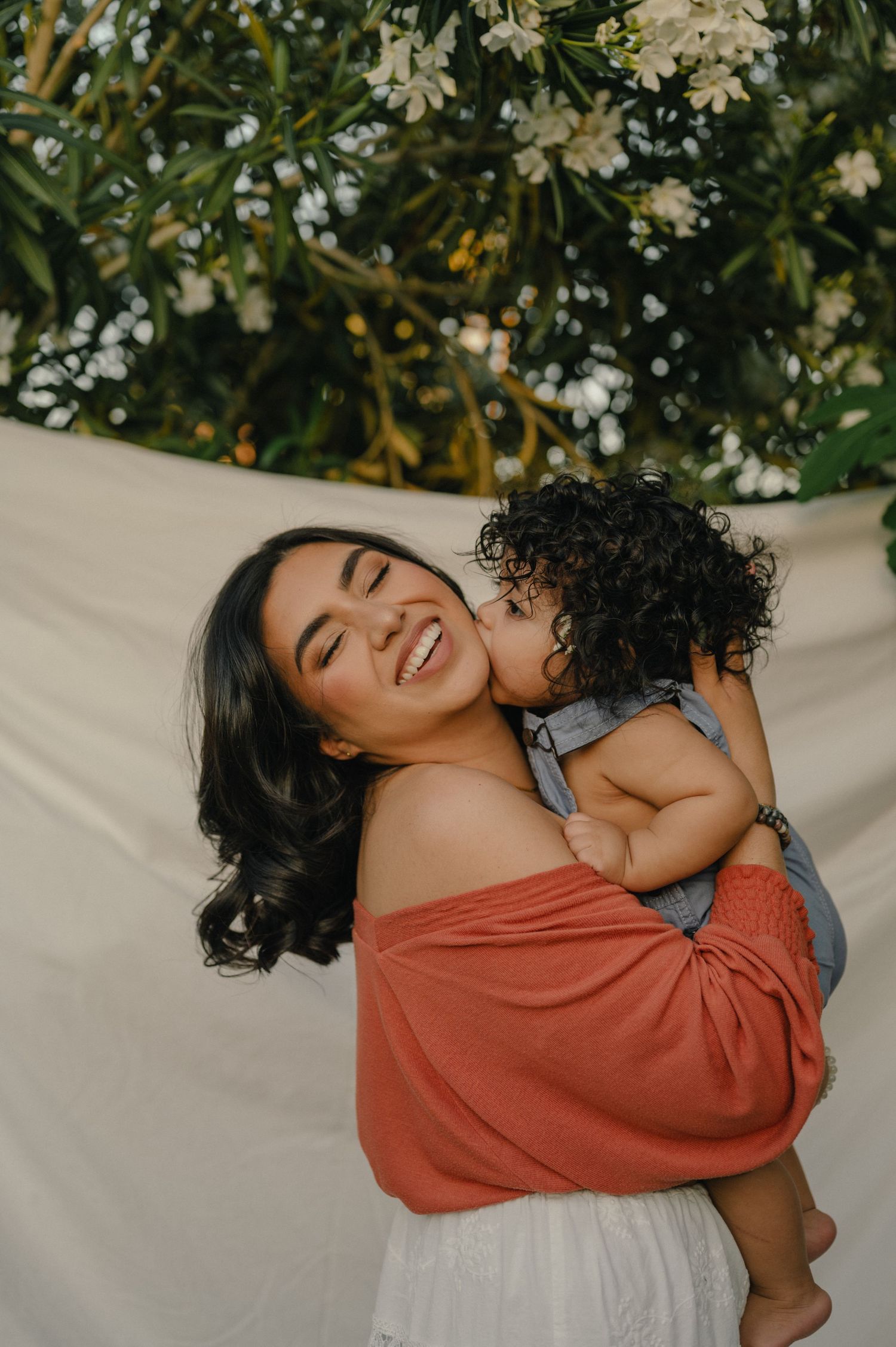 Smiling mother in rust off-shoulder top joyfully holding her curly-haired baby near blooming flowers.