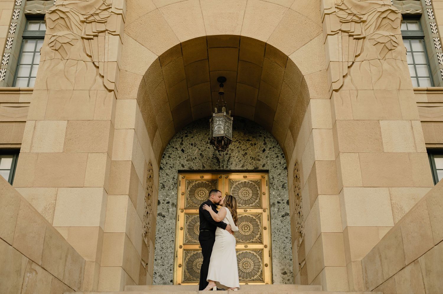 Couple embracing in front of ornate Art Deco building entrance with golden doors and stone arch.