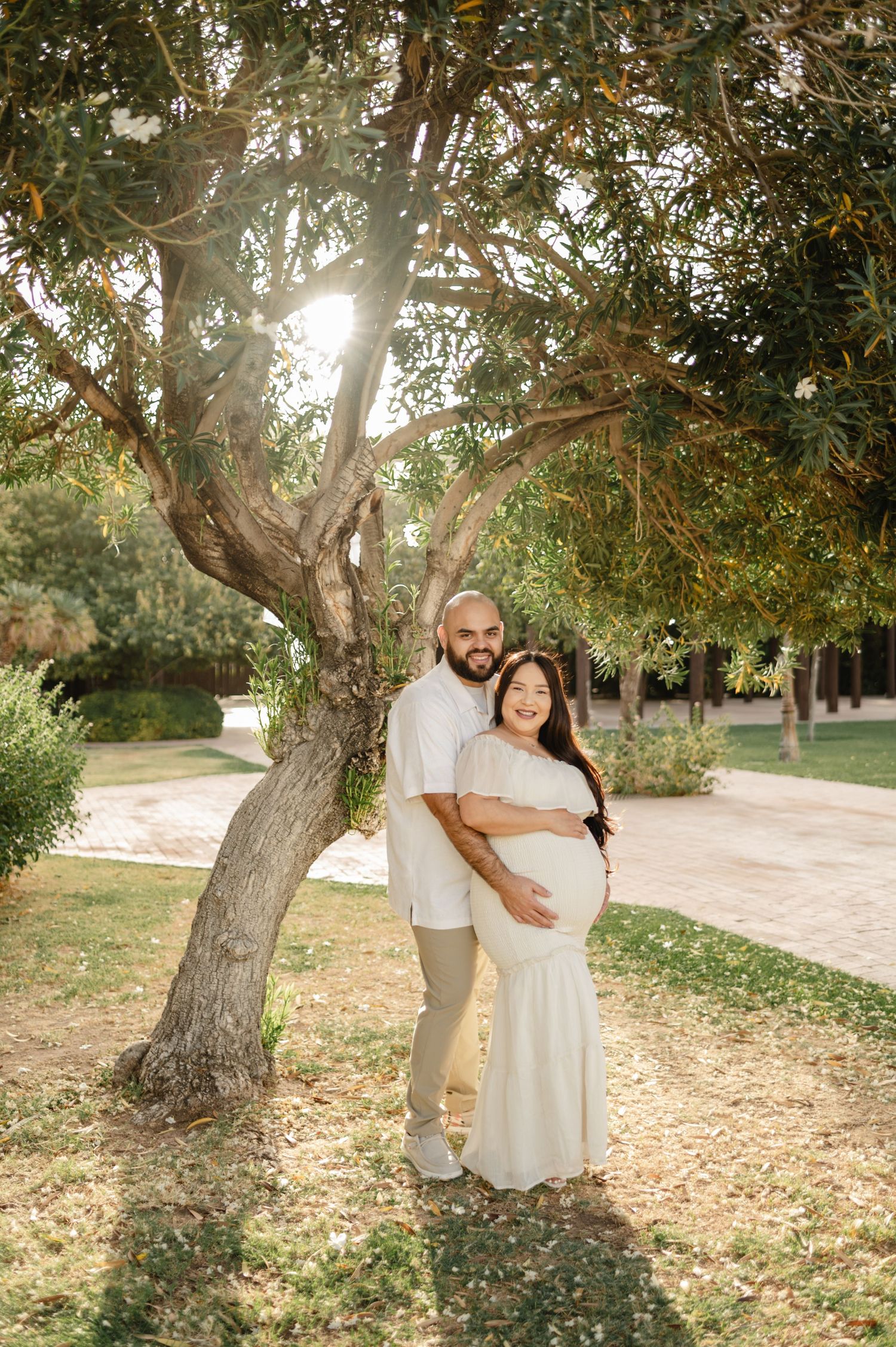 Couple poses under a sunlit tree in a park, woman in white maternity dress cradling her bump, man embracing her from behind.