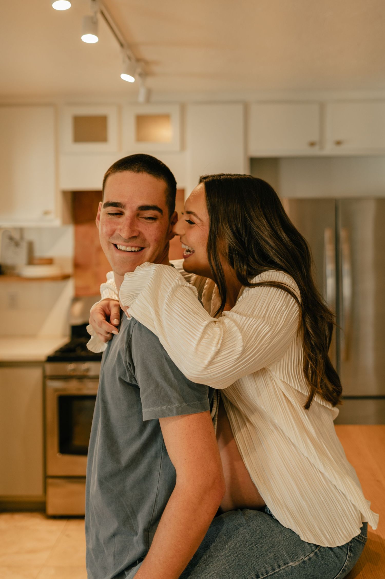 Happy couple laughing together in bright kitchen, woman riding on mans back.