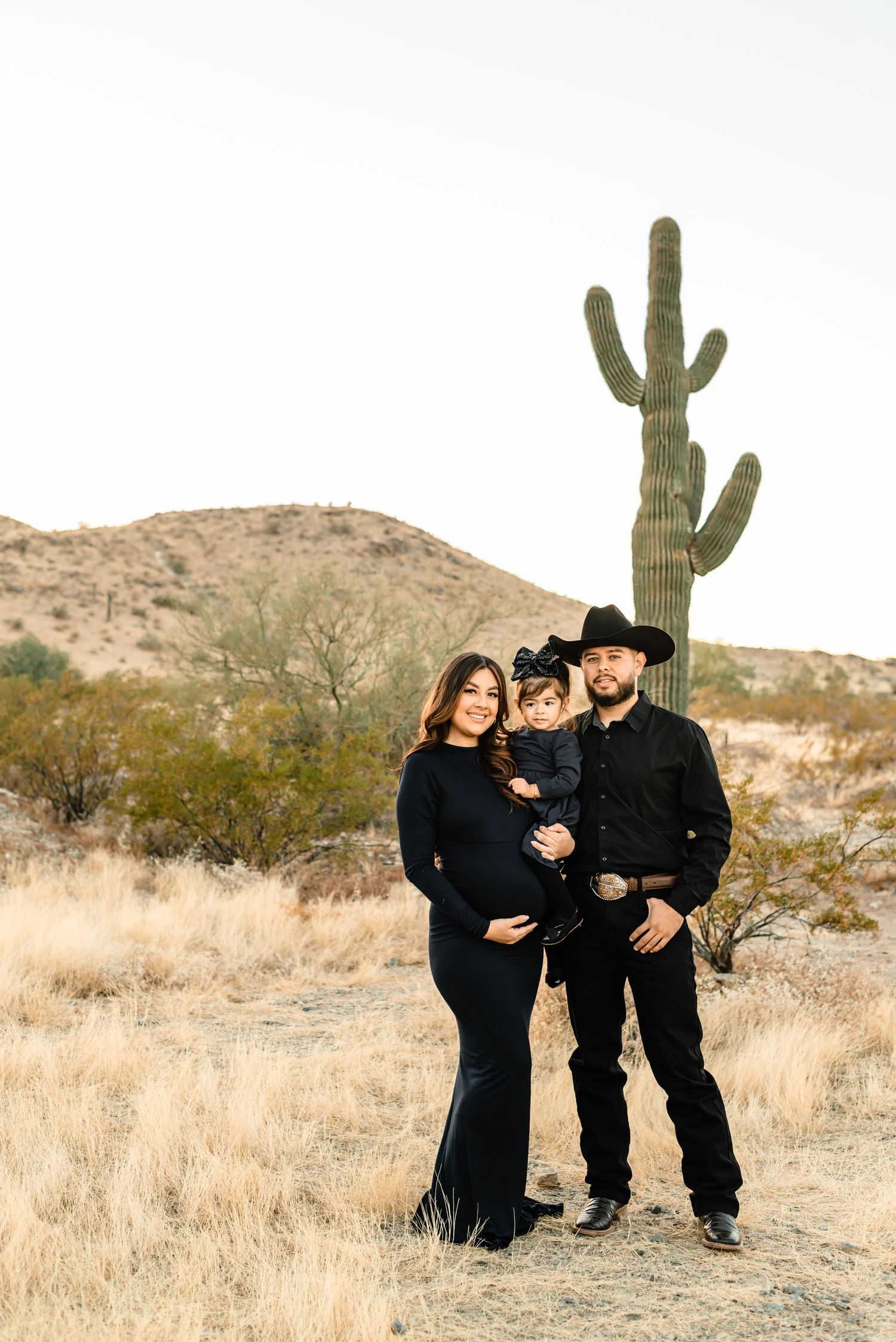 Family of three in black outfits poses before a saguaro cactus in the Arizona desert, maternity photo session.