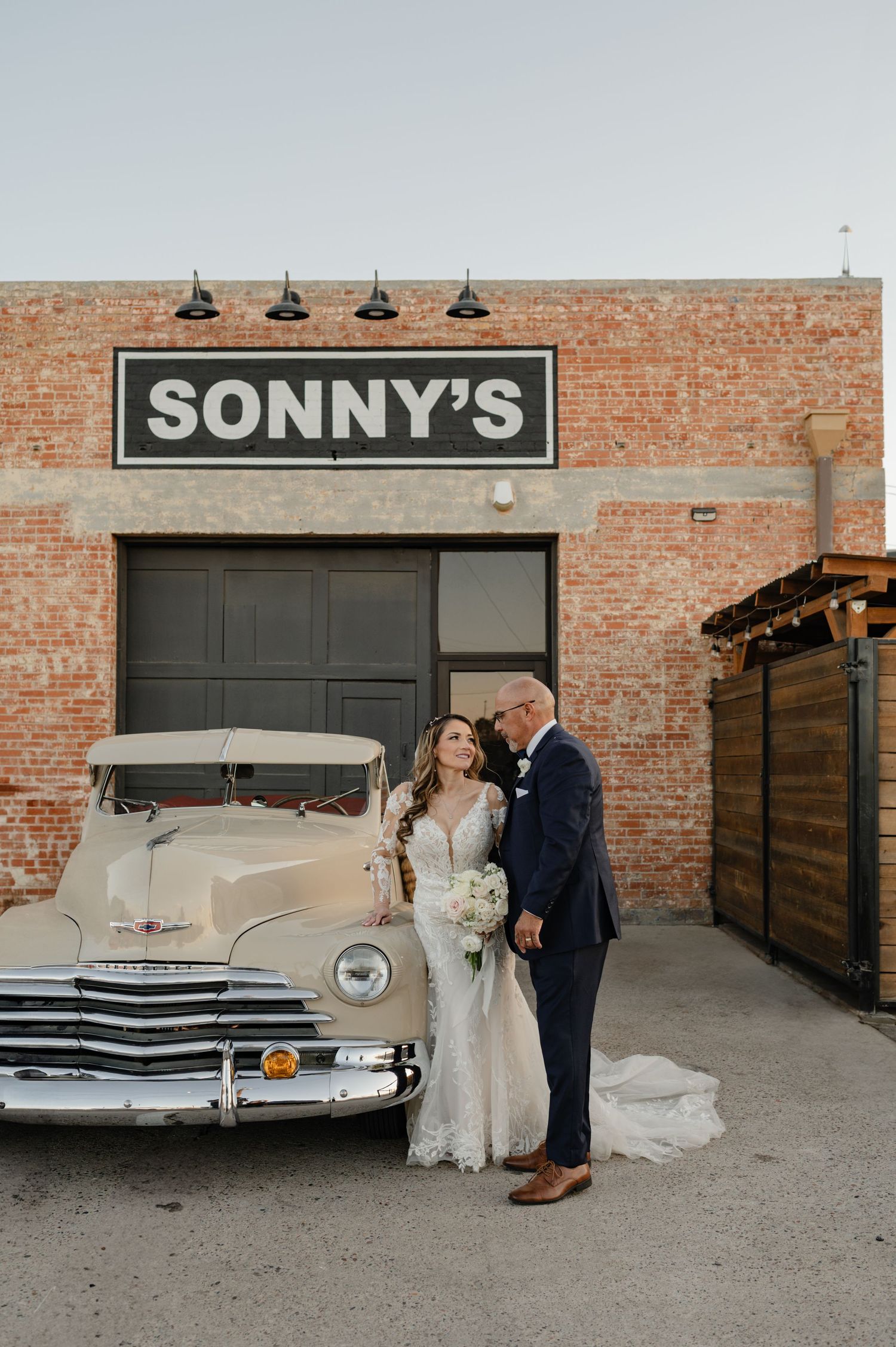 Bride and groom posing beside vintage car outside Sonny’s, a downtown Phoenix wedding venue with brick backdrop.