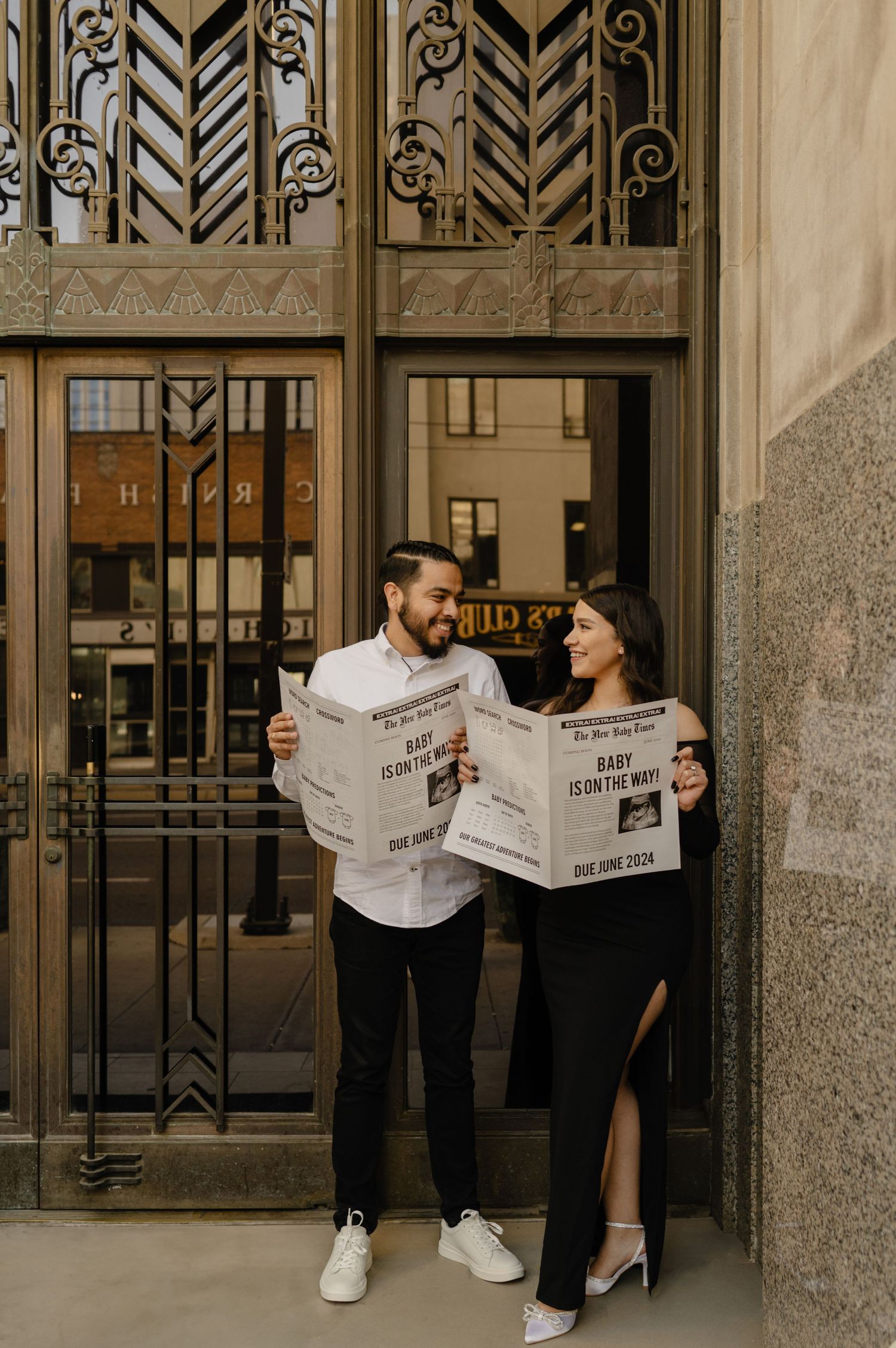 couple laughing at each other holding baby is on the way newspapers in front of an art deco doorway, downtown phoenix pregnancy announcement