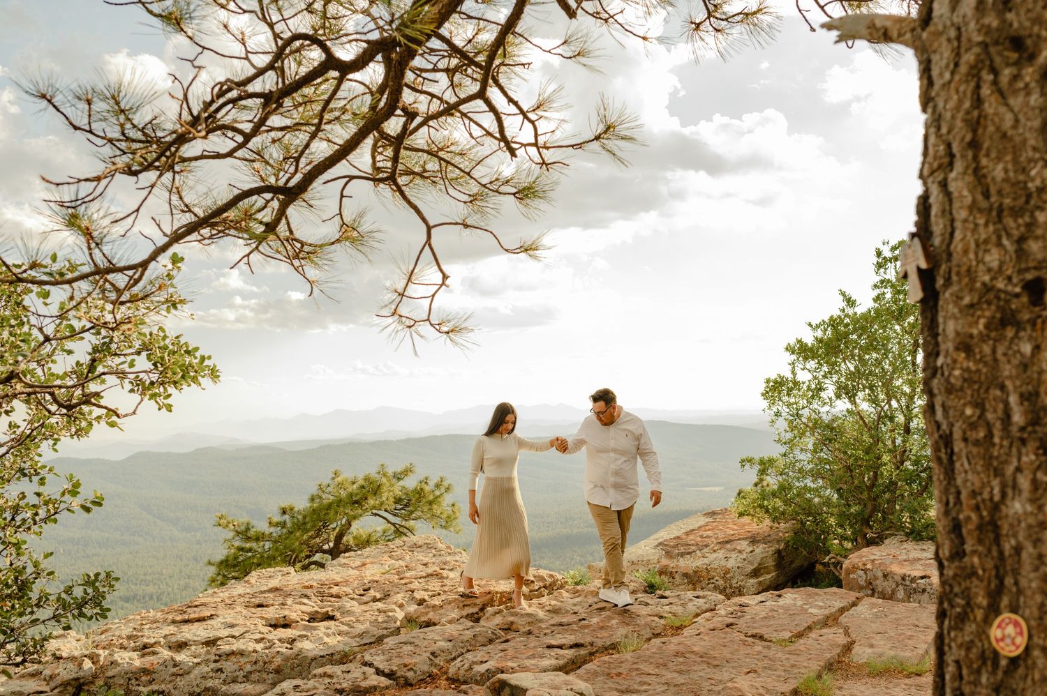 Couple walks hand in hand on rocky cliffside surrounded by pine trees and mountain views.