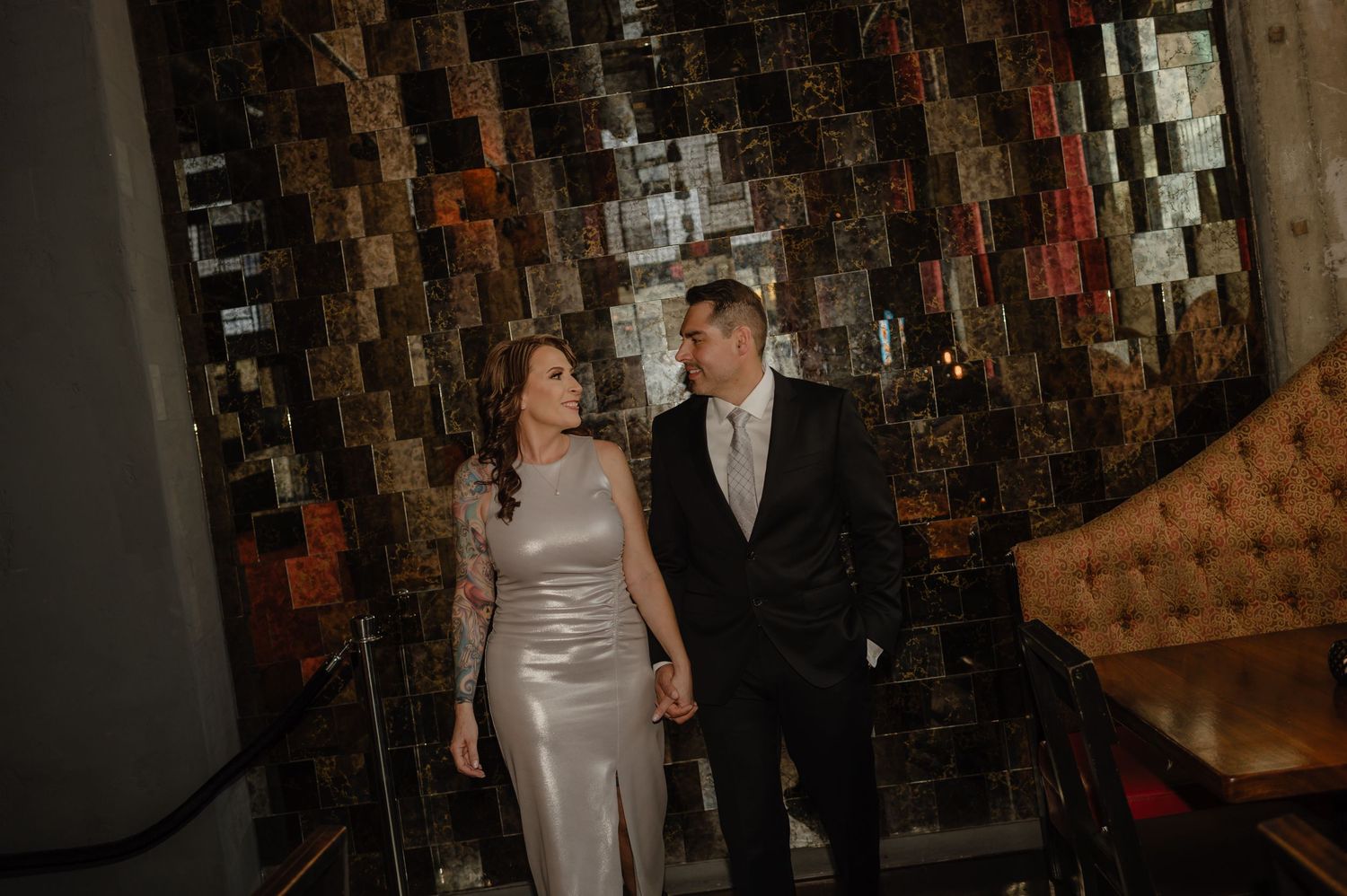 Elegant couple in formal attire holding hands on stairs before a decorative mirrored wall at an upscale venue.