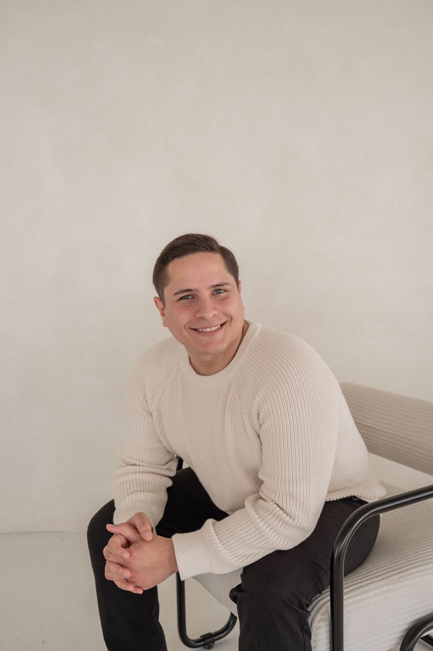 Man sitting on a stool in a studio space, smiling — formal portrait of one half of the creative duo.