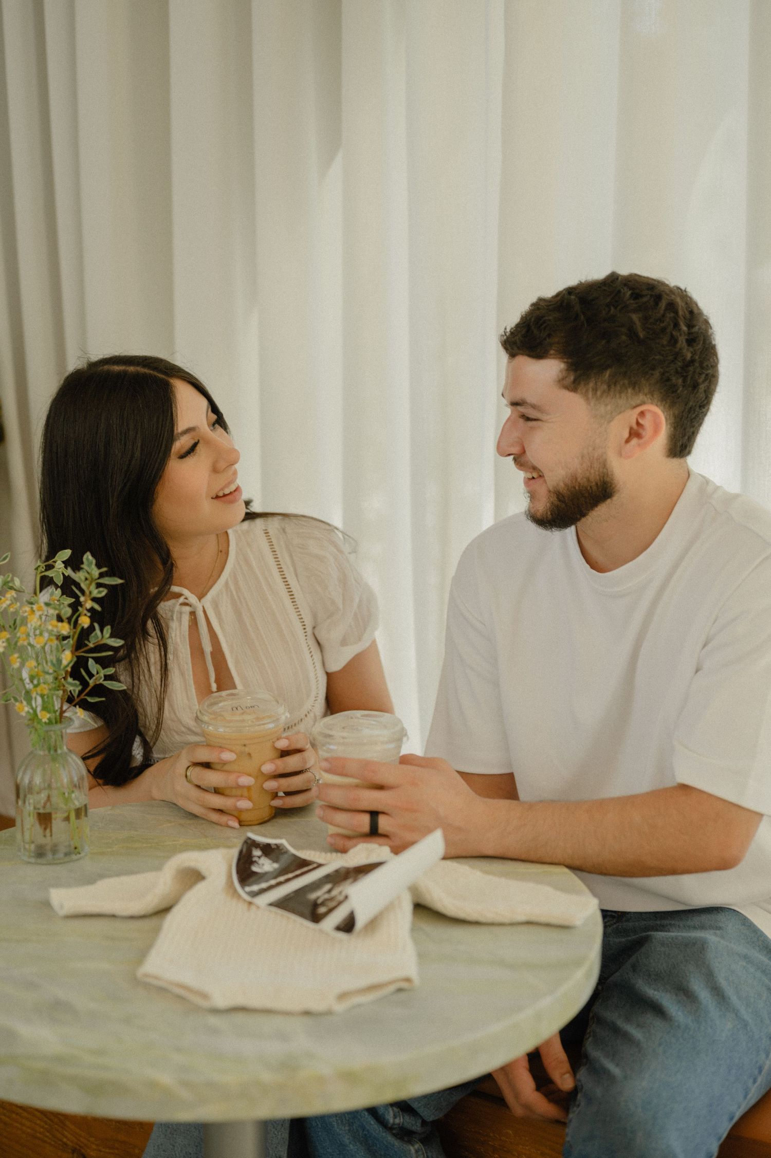 couple smiling at each other over iced coffees with an ultrasound and knit baby outfit on the table, cafe pregnancy announcement