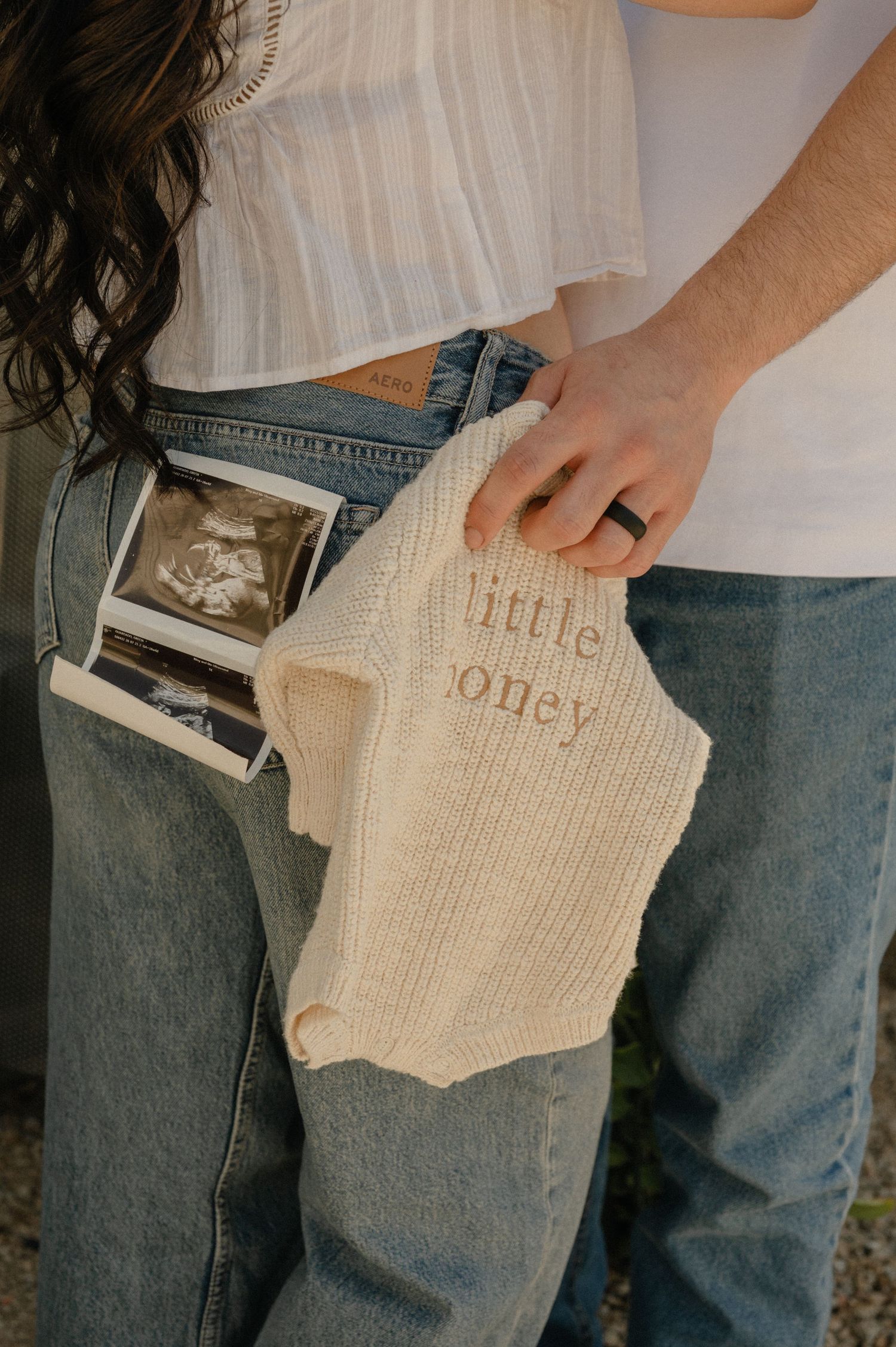 ultrasound photo tucked in a back jeans pocket while a partner holds a knit baby outfit, pregnancy announcement photographer glendale az