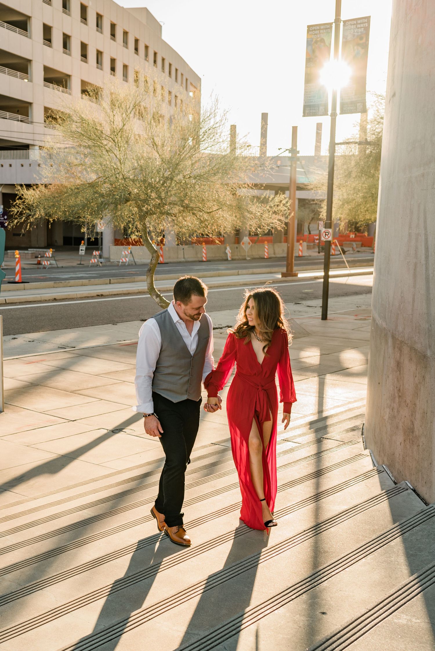 Couple holding hands walking in urban setting, woman in red dress, man in vest, golden sunlight casting long shadows.