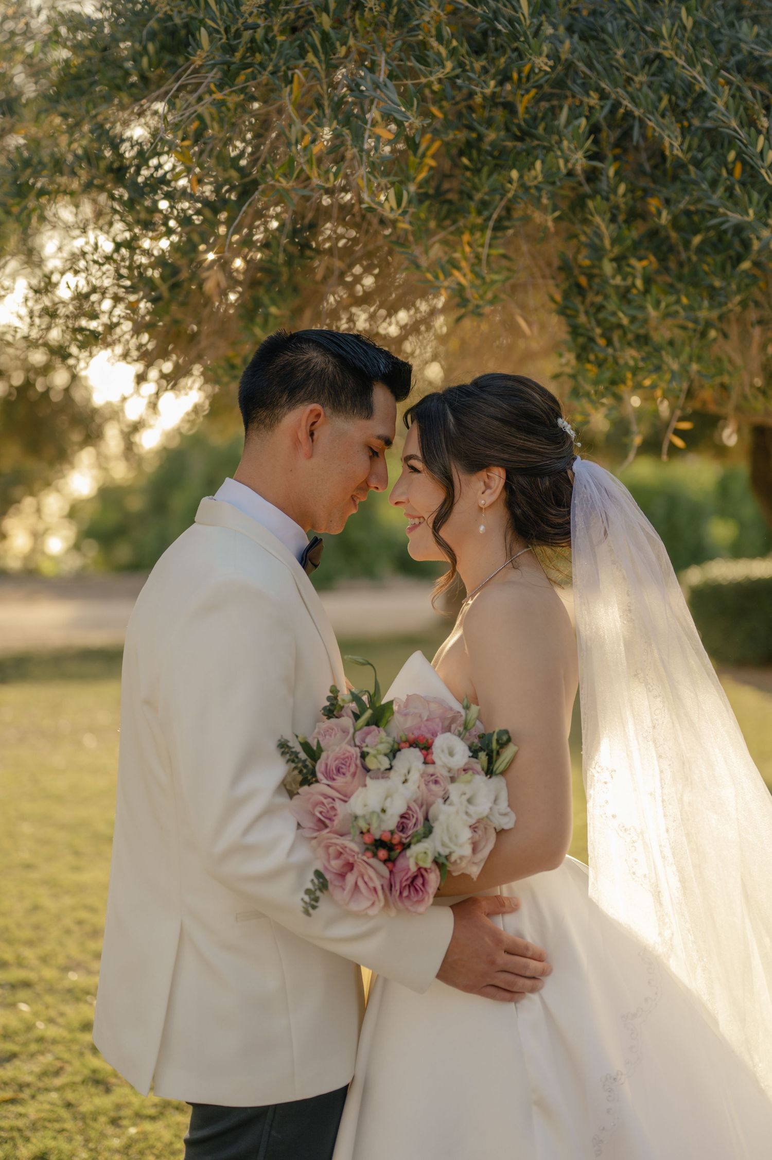 Bride and groom face each other lovingly, bride holding soft pink bouquet beneath tree.