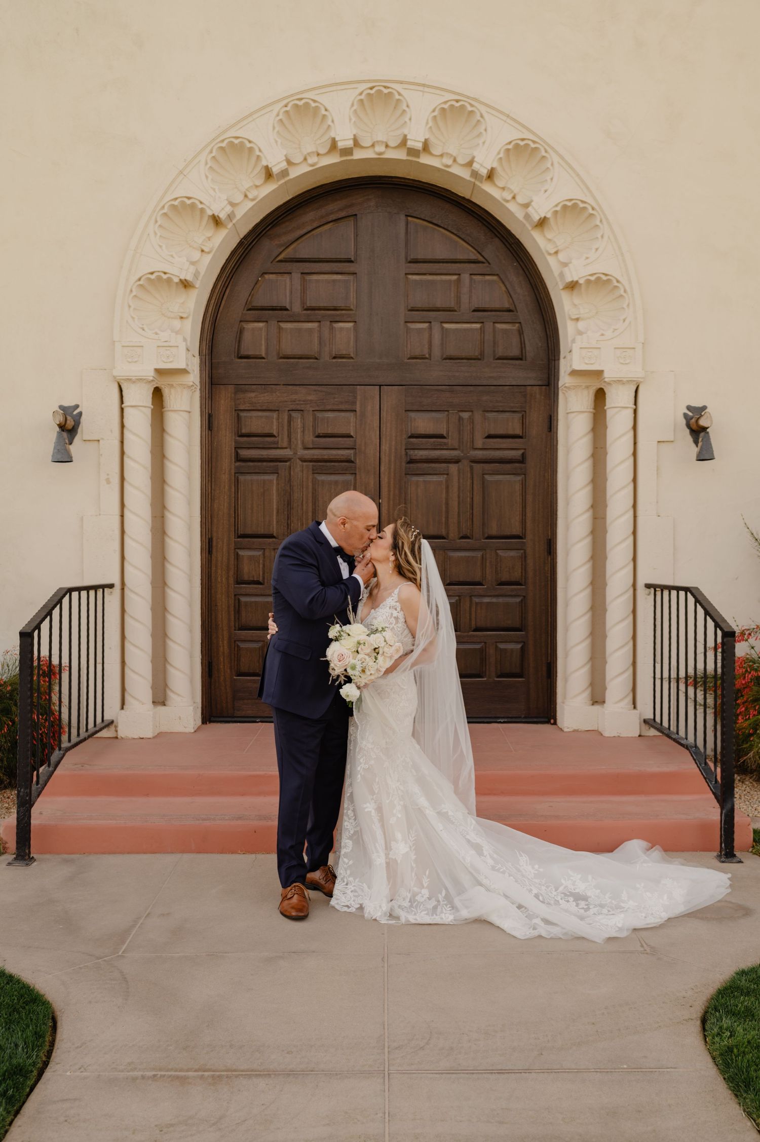 Bride and groom kiss on steps of historic church with arched wooden doors and elegant cream-colored architecture.