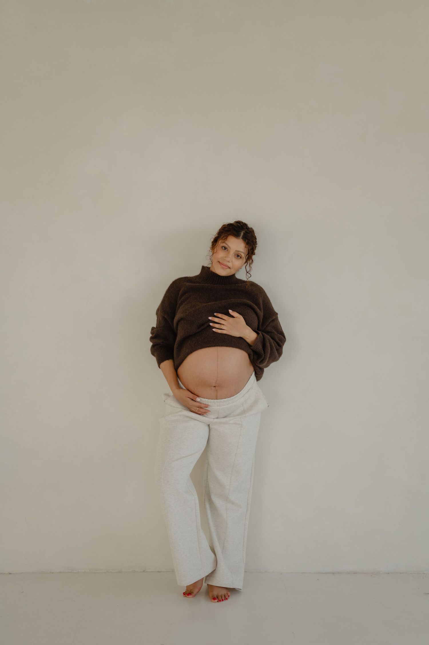 pregnant woman in a brown sweater and white pants leans against a studio wall during a phoenix maternity session at refine studios