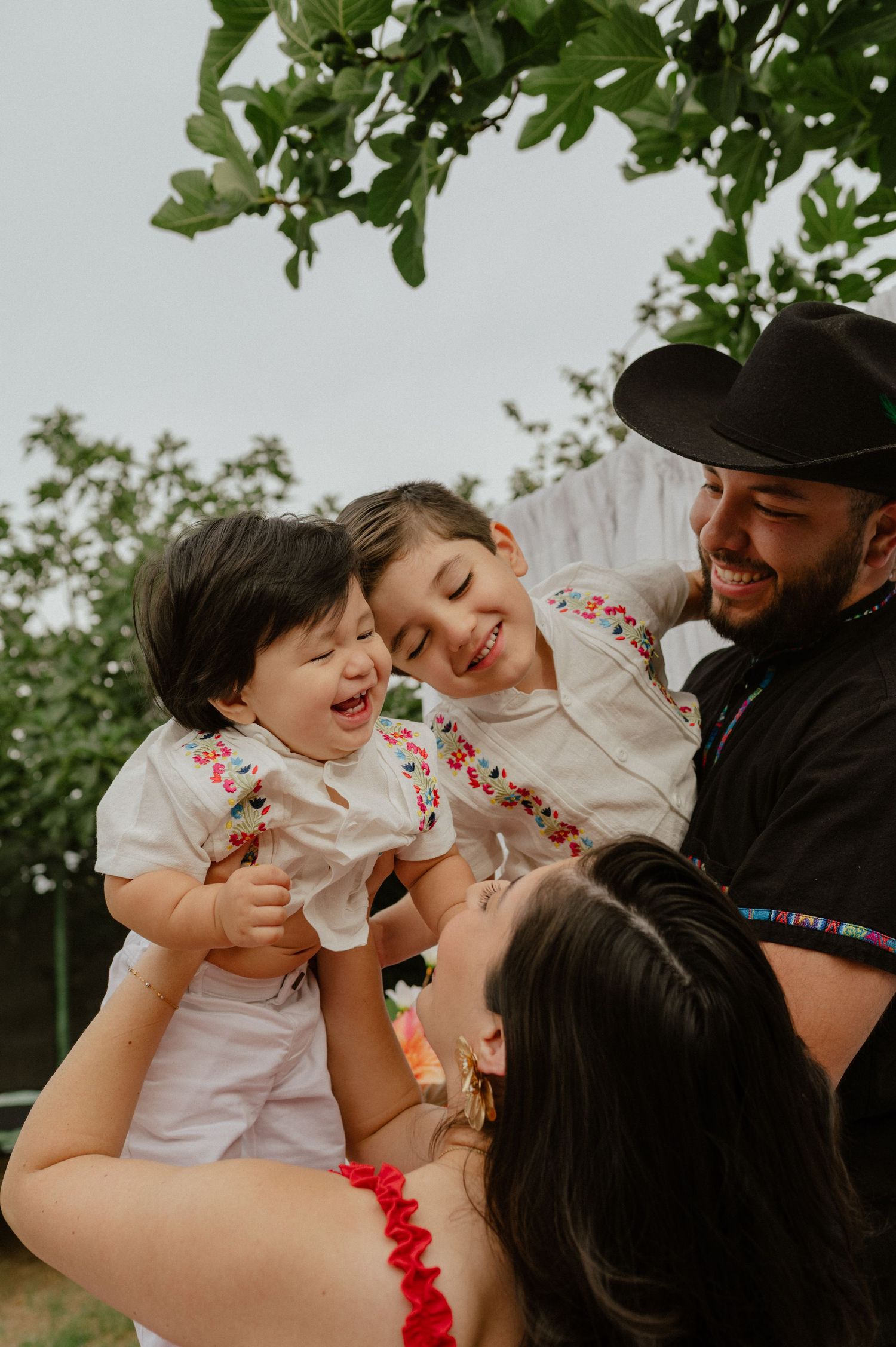 Joyful family with father in black cowboy hat holding two laughing children in embroidered white outfits.