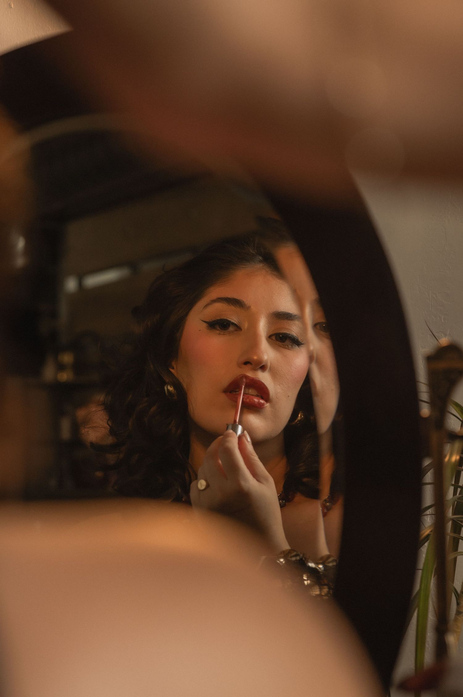 Woman applies lipstick while looking into a round mirror in a softly lit salon setting.