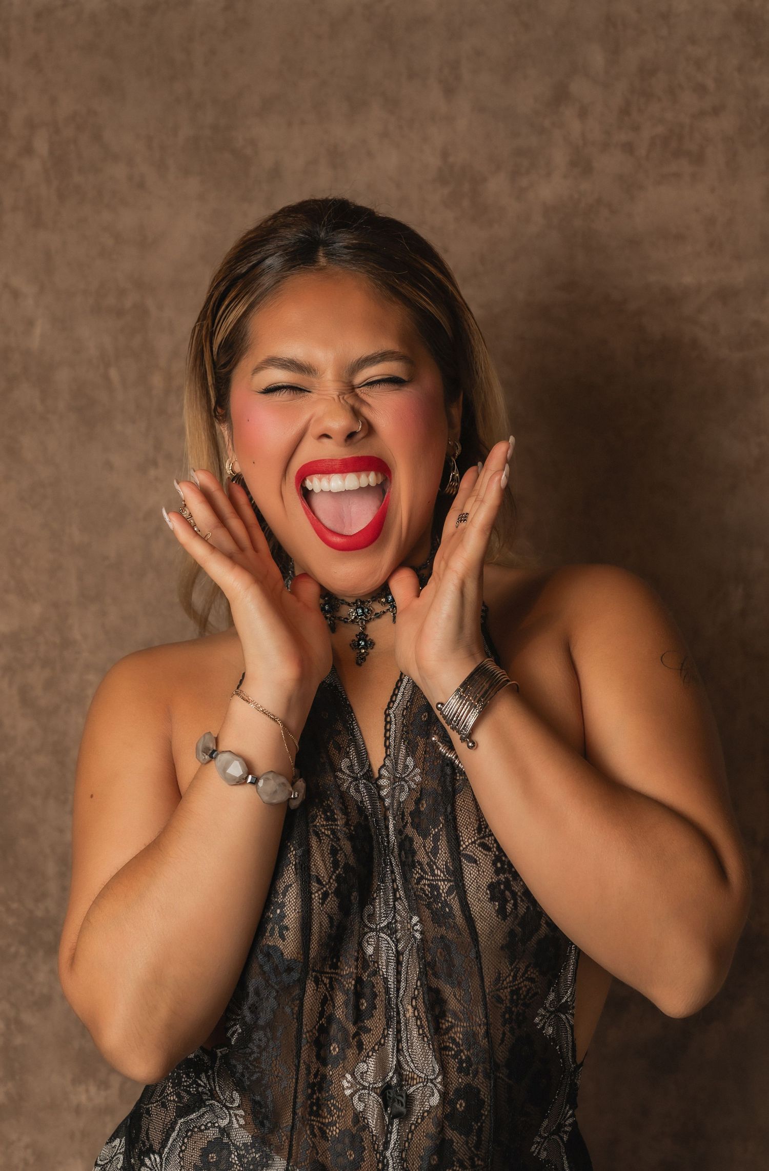 Woman joyfully screams with red lipstick and expressive hands in front of brown backdrop.