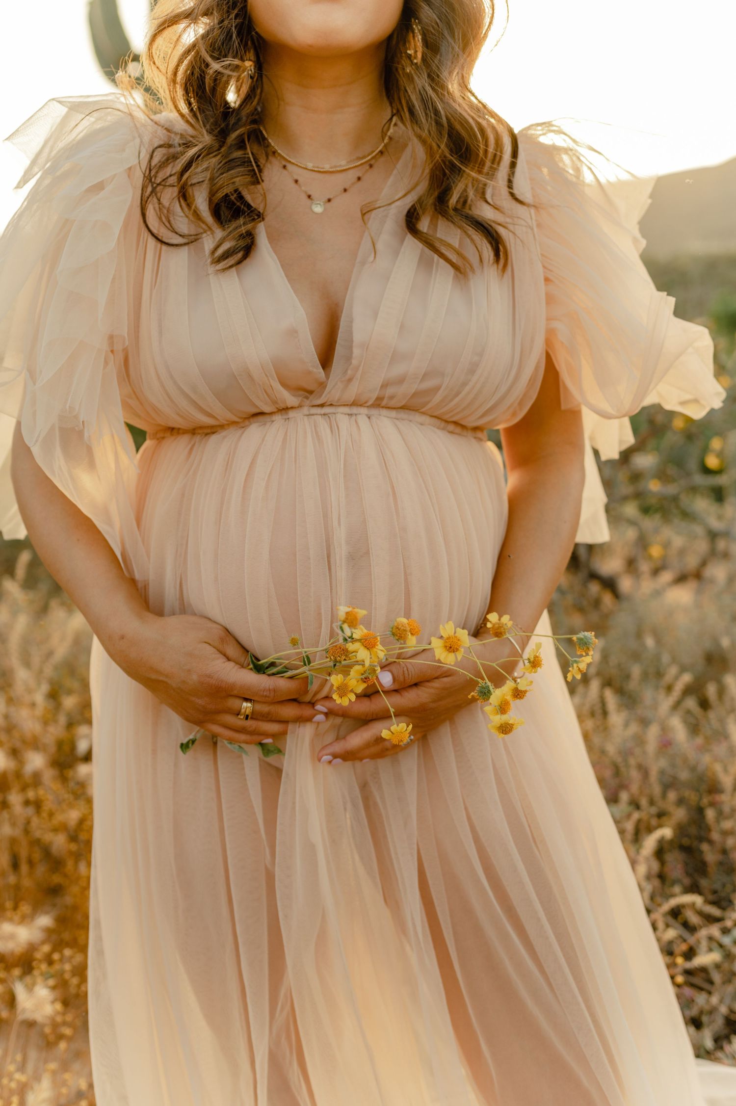 Pregnant woman in peach tulle dress holding yellow wildflowers outdoors.