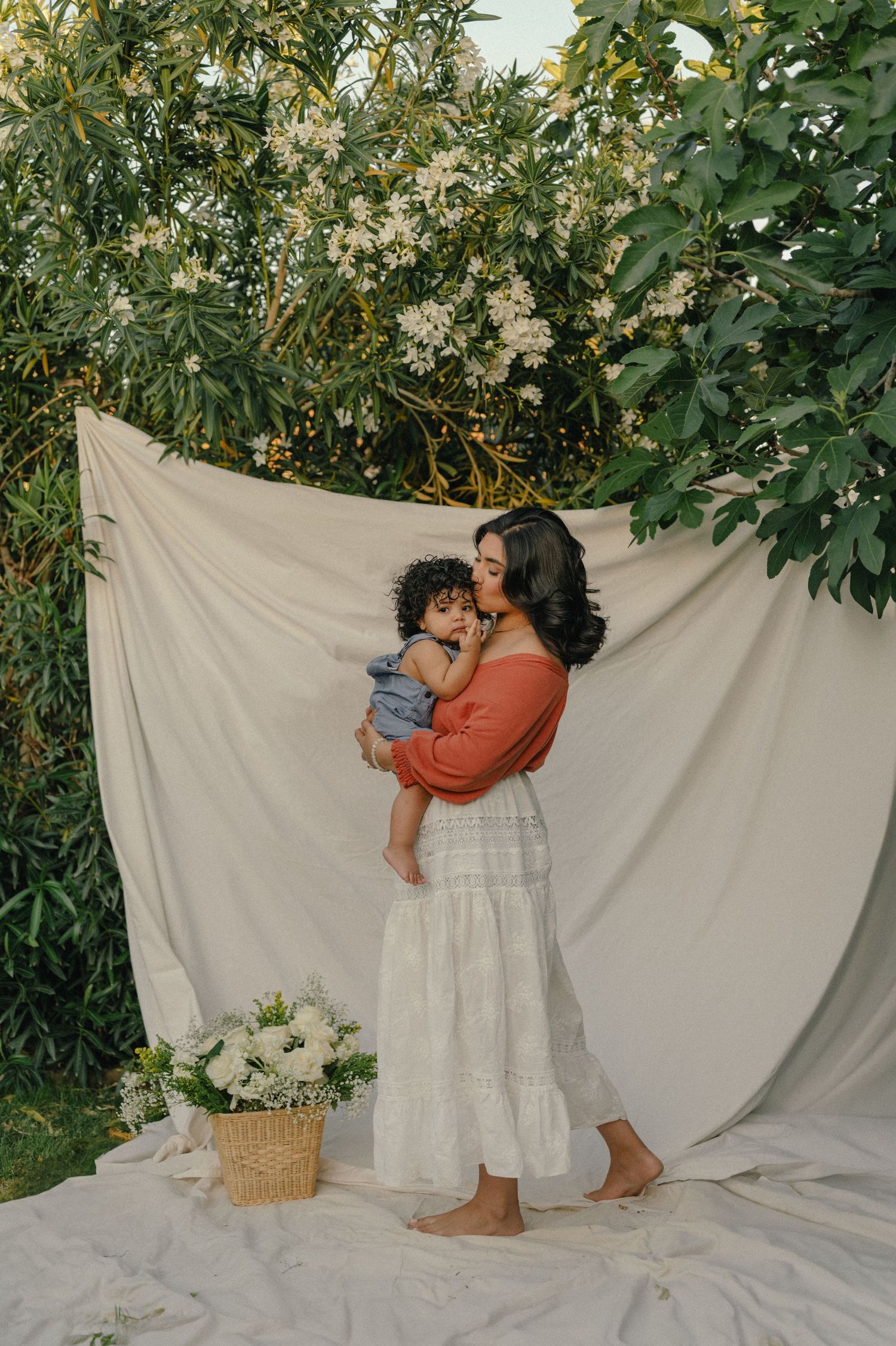 Mother in orange top and white skirt kissing baby outdoors against white fabric backdrop with flowers.