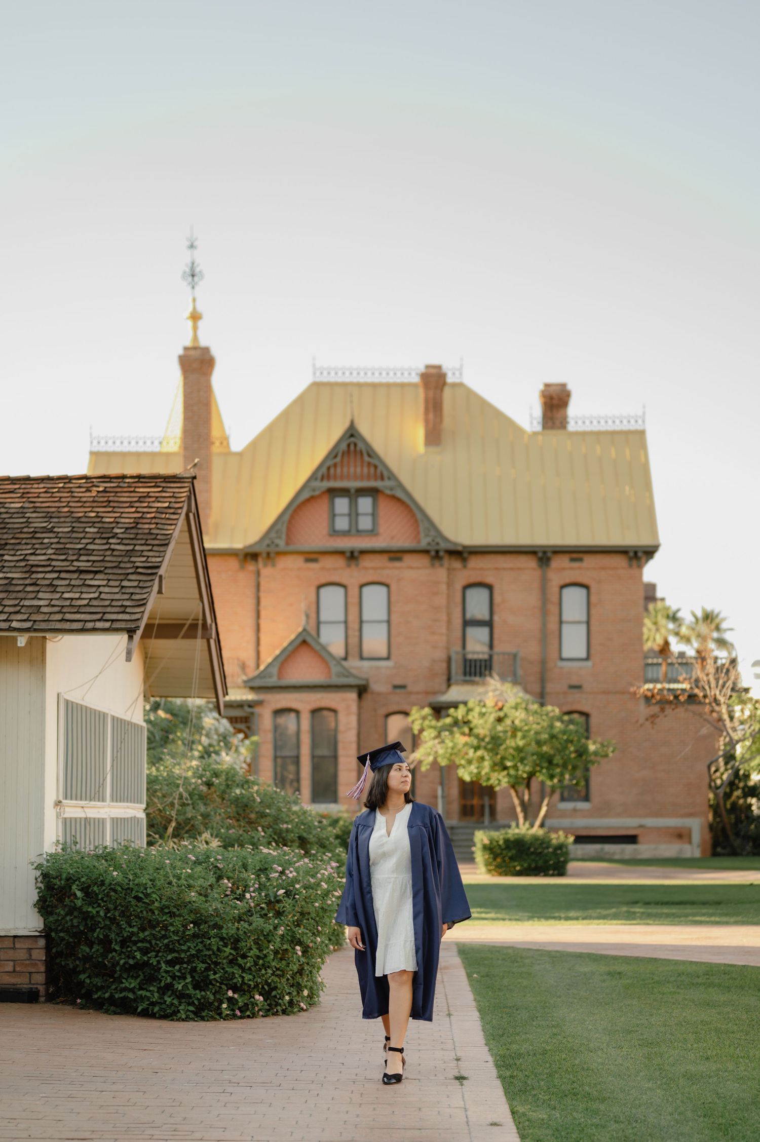Graduate in navy cap and gown walks on brick path before historic red brick Victorian building on sunny day.