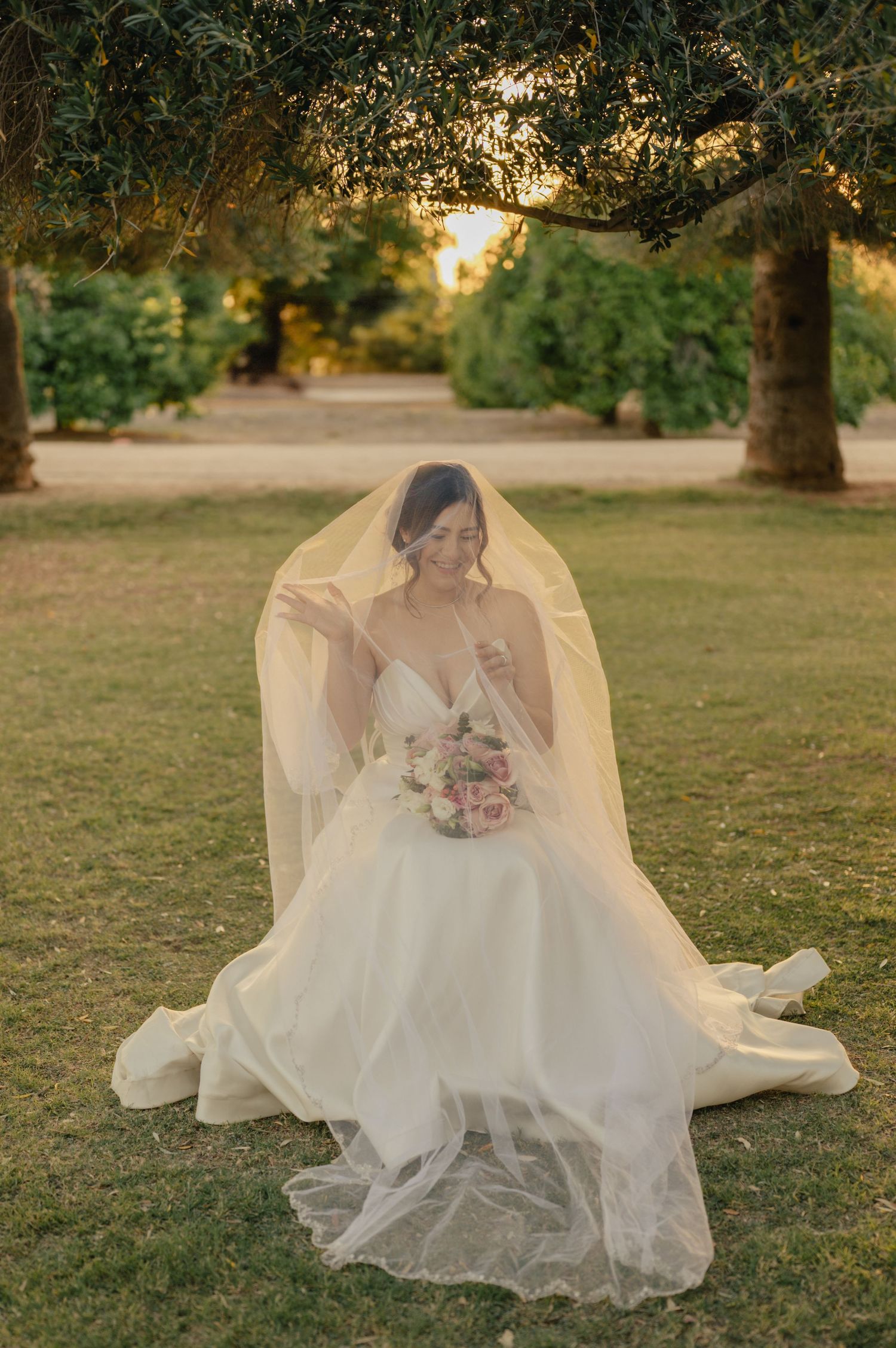 Bride kneels in grass at golden hour, holding bouquet and veil draped around her.