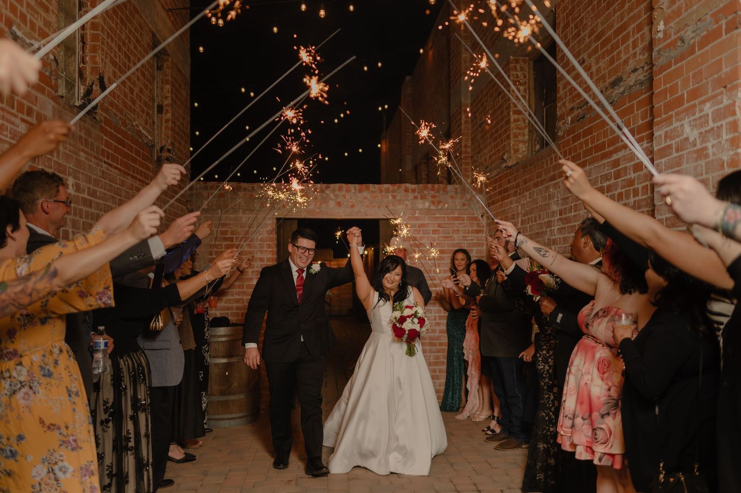 Newlyweds walk through sparkler tunnel surrounded by cheering guests in brick alleyway.