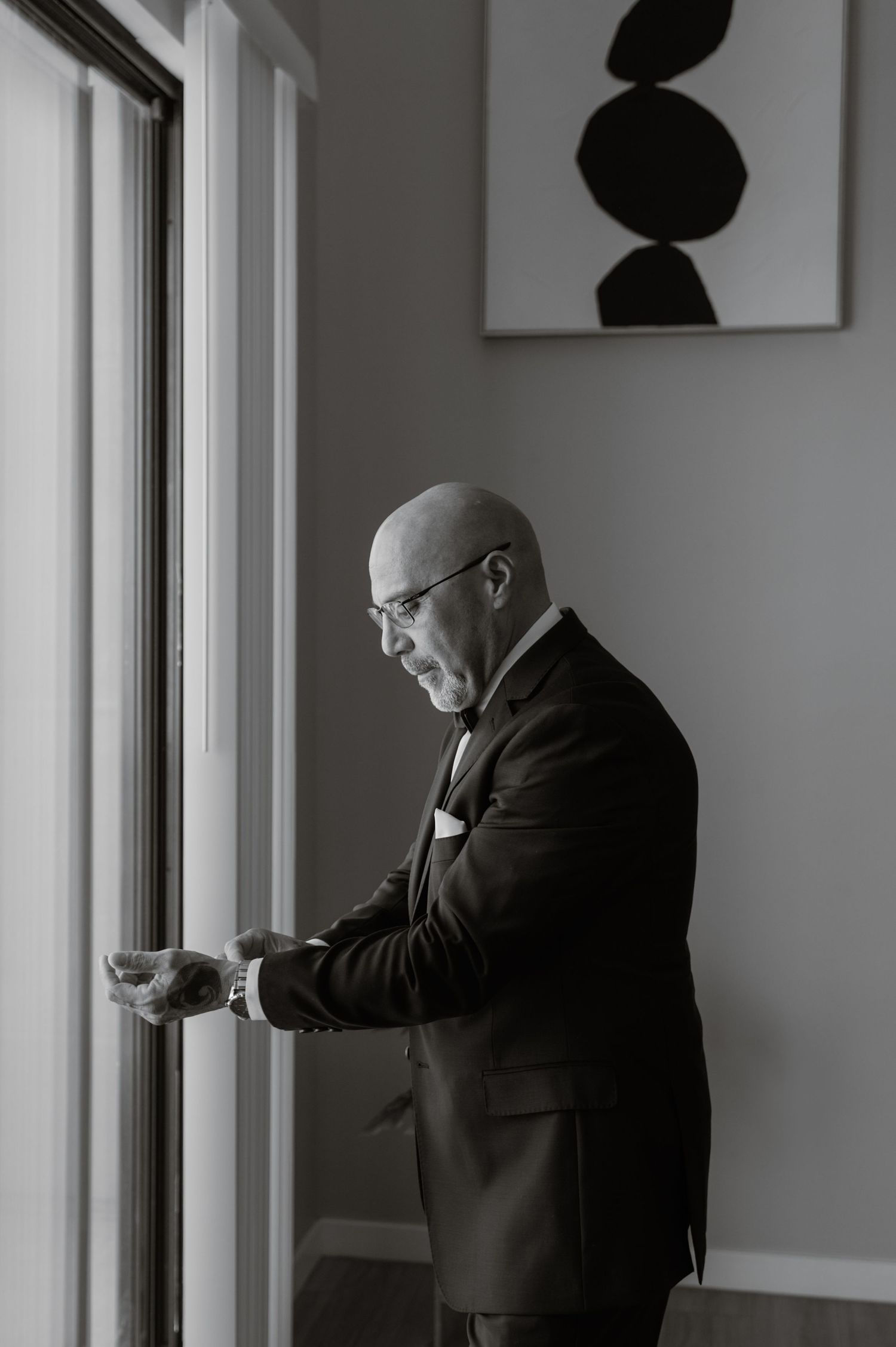 Groom in black suit adjusting cufflinks while standing at a window in soft natural light, quiet moment pre-ceremony.